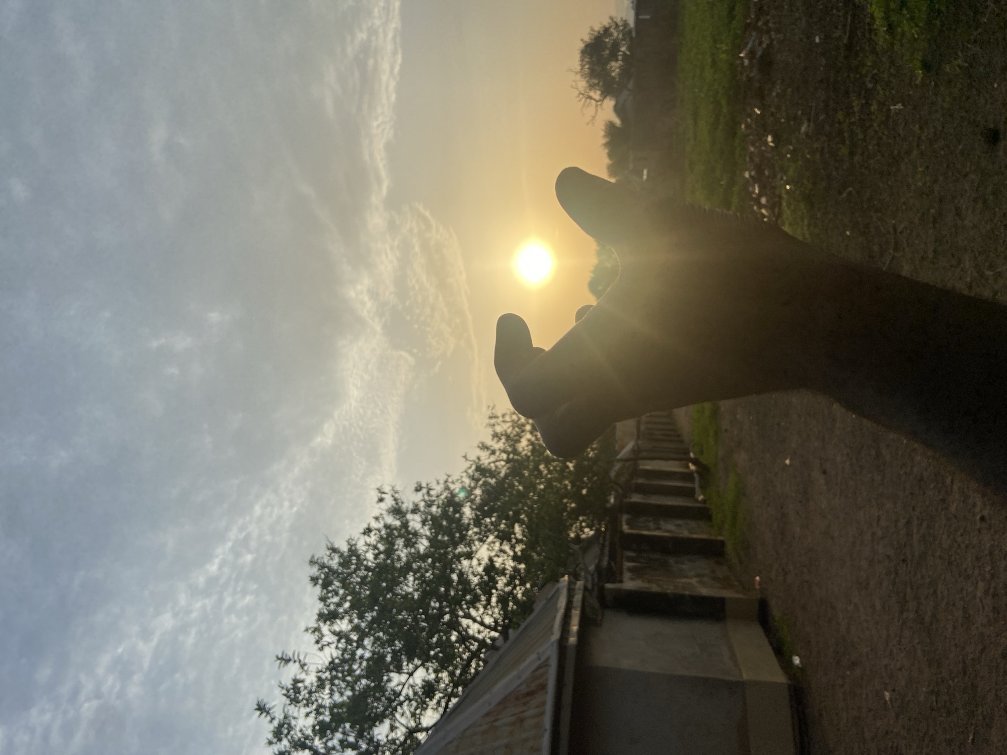 A low-angle photograph shows a hand reaching toward the sun in Mararaba, Nigeria. The sun is bright, partially obscured by the fingers of the hand. The sky has scattered clouds. In the foreground, there is a building with a metal roof and a staircase along a wall. A tree is near the building. Grass and foliage are visible in the background, along with a path or road. The image has a warm, golden light.