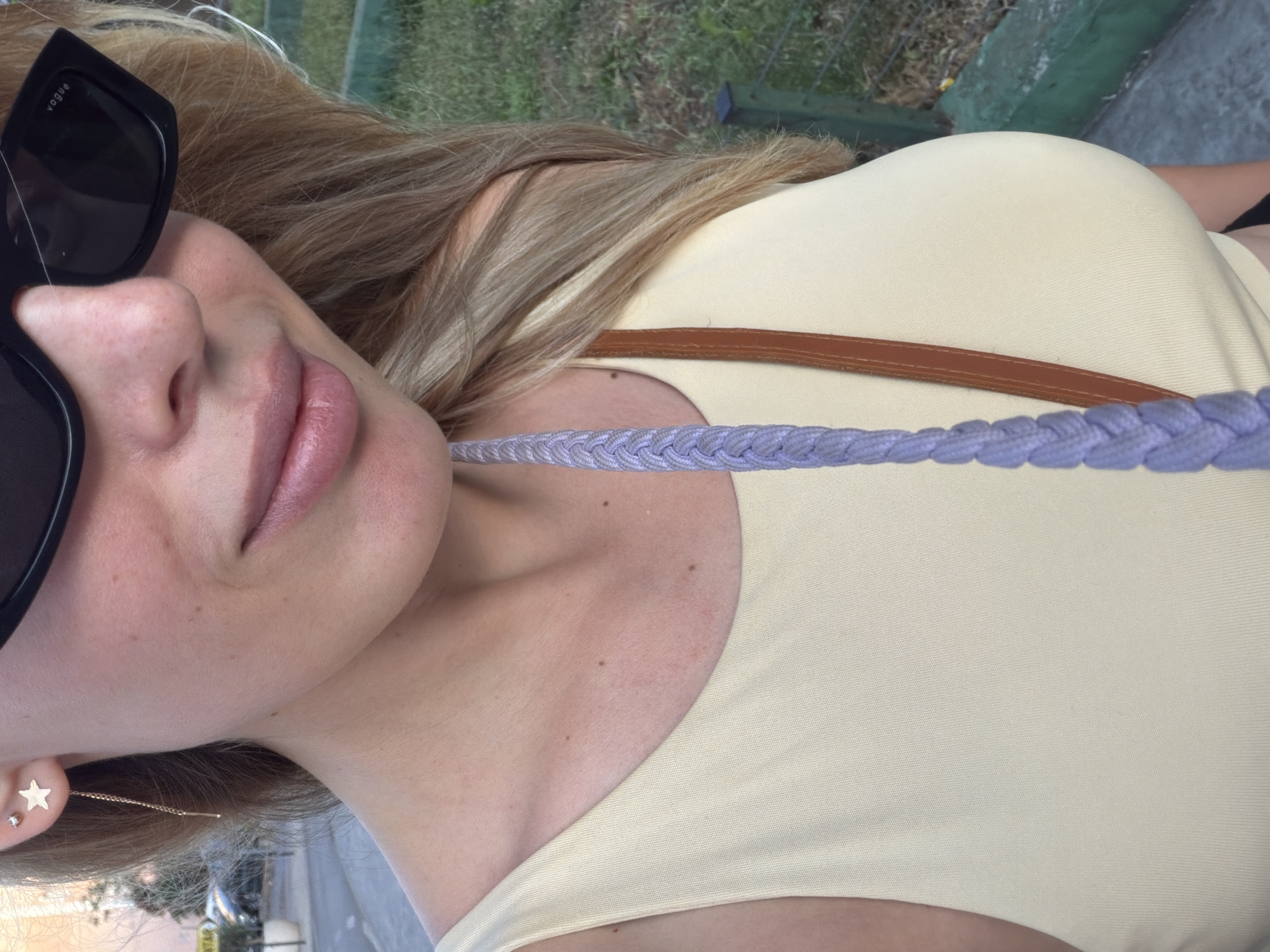 The image shows a close-up of a woman's upper body, from the neck up, as she stands outdoors. She is wearing a light yellow sleeveless top and sunglasses. A thin brown strap from a bag crosses her chest. Her blonde hair is visible, falling over her shoulders. A braided cord, appearing to be lavender in color, hangs down the front of her. A star-shaped earring is visible on her left ear. The background consists of greenery and a paved area, suggesting an outdoor urban or park setting. The context suggests the image was taken in Budva, Montenegro, though no specific landmarks confirming this location are visible within the frame.