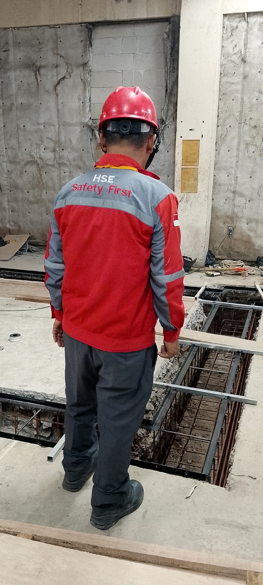 A full shot, rear view, shows a person wearing a red and gray work jacket with "HSE Safety First" written on the back, a red hard hat, and dark pants. The person is standing on a concrete surface, looking down at a trench with rebar reinforcement. The environment appears to be an indoor construction site, possibly a building under renovation or construction, in Depok, Indonesia. The background shows unfinished walls, possibly made of concrete blocks, and scattered construction materials like wood planks and electrical cords.