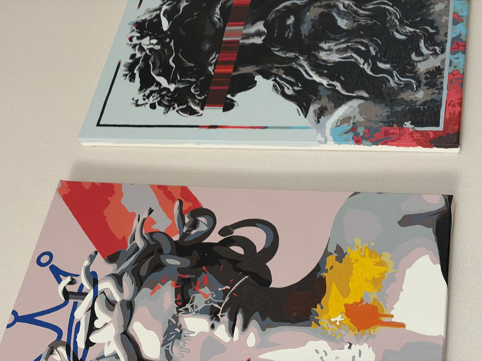 The image displays two contemporary, stylized paintings arranged vertically against a plain, light-colored wall, likely indoors. Both artworks feature abstract interpretations of classical busts or heads, characterized by a pop-art or paint-by-numbers aesthetic with distinct blocks of color and outlines.
The upper painting presents a head with flowing, dark and grey hair-like elements, possibly resembling a male bust or a mythical figure, set against a light blue background. Intersecting the figure are bold red and black geometric lines and shapes. The edges of this canvas show additional red and blue brushstrokes, giving it a somewhat unfinished or raw aesthetic on the sides.
The lower painting depicts a profile view of a head, possibly Medusa, featuring prominent dark, coiled, and serpentine hair against a light pink or lavender background. This artwork incorporates vibrant accents of bright yellow, orange, and red, particularly around the face and hair. A distinctive blue, loop-like graphic element is visible on the left side of this painting, adding to its modern, graphic style. Near the bottom center of the lower painting, faint cursive text, possibly a signature like "Mirallo," can be discerned.
Both canvases have visible edges, suggesting they are either leaning against the wall or freshly mounted. The overall impression is one of modern artistic interpretations that blend classical subject matter with a contemporary, graphic sensibility.
