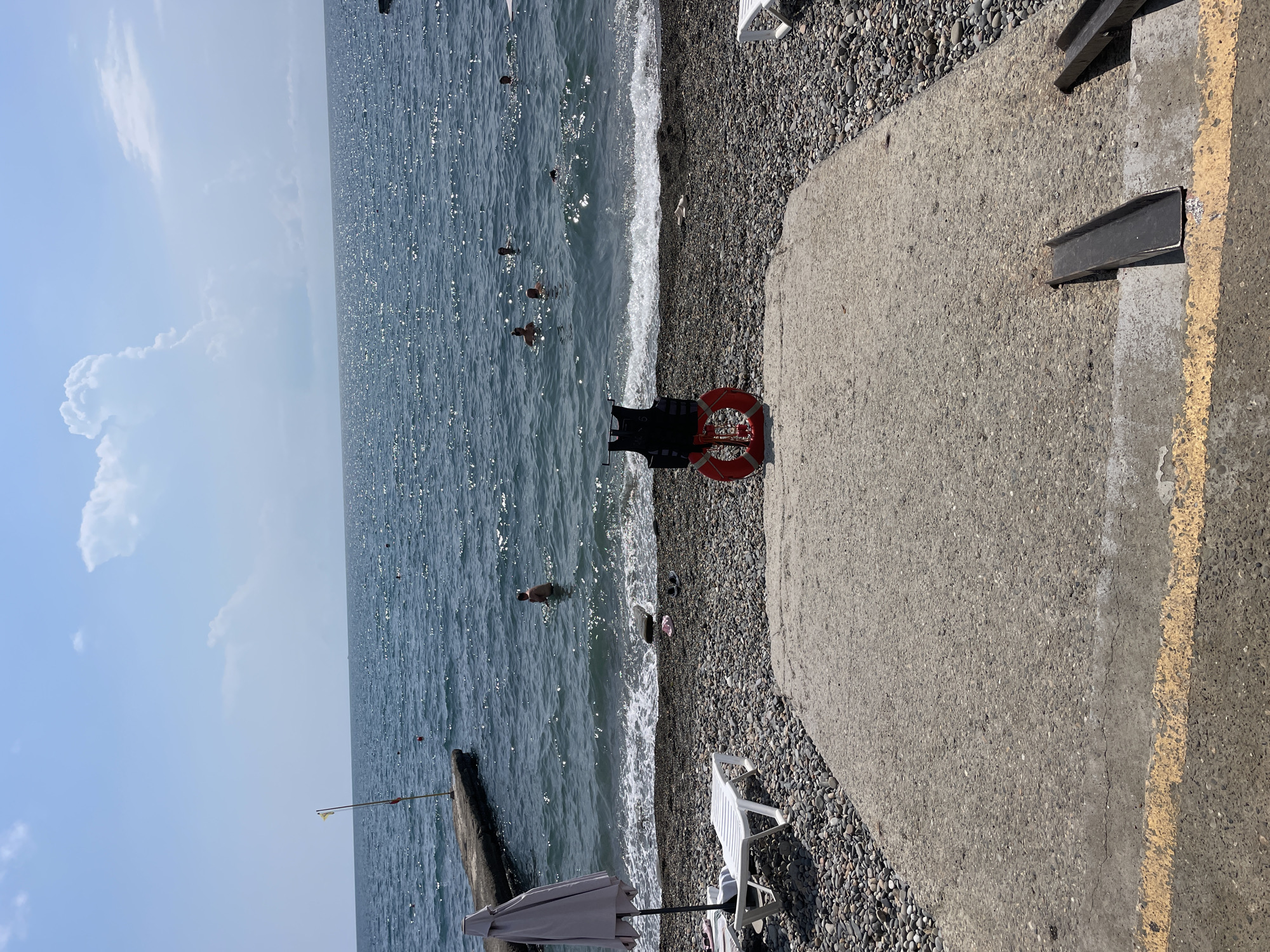 The image depicts a sunny day at a pebble beach in Sochi, Russia. The sea is calm with gentle waves rolling onto the shore. In the water, several people are swimming and enjoying the ocean. On the beach, there are two white lounge chairs and a folded beach umbrella, suggesting a relaxed atmosphere. In the foreground, a black life vest and a red lifebuoy are placed on a concrete ramp leading down to the beach, possibly for safety or to indicate a designated swimming area. A concrete pier with a yellow flag extends into the sea on the left side of the image. The sky is a clear blue with a few scattered white clouds.