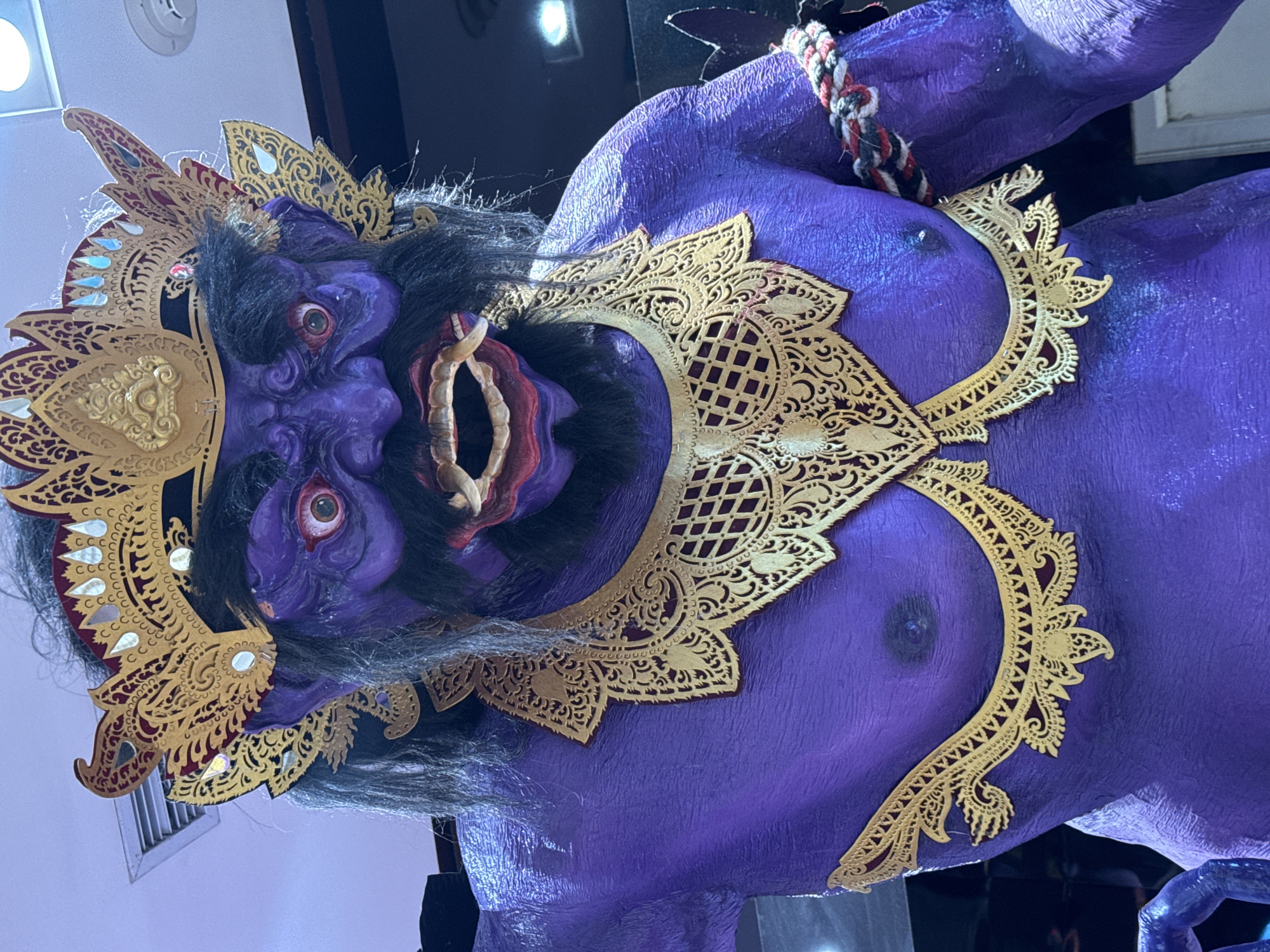 A close-up, somewhat vertically oriented photograph depicts a striking purple statue adorned with intricate golden ornamentation. The statue is likely a Balinese Barong, a protective spirit often depicted in dance and performances in Bali, Indonesia. The figure has a fierce expression, with bulging red eyes, sharp teeth, and black hair and beard. Golden decorative elements surround its head and chest, adding to its regal and imposing appearance. The statue's purple skin is textured, possibly to emulate a natural or mythical surface. The presence of a bare chest suggests this statue is of a male deity. The backdrop is indistinct, but hints of lighting suggest that this statue is indoors. Without more information, I cannot say for sure that this is located in Kuta, Indonesia.