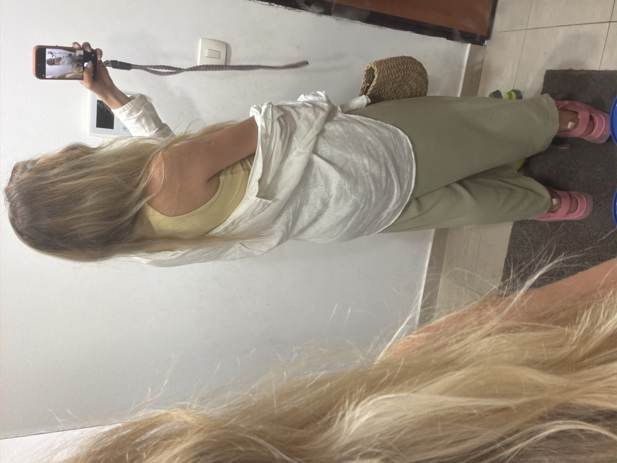 The image shows a person holding a smartphone in their left hand, taking a selfie. The person has long, blonde hair and is wearing a white shirt and light green pants. They are also wearing pink slides. In the background, there is a wall with a light switch and a small woven basket. The scene appears to be indoors, possibly in a home or hotel room. The lighting suggests it might be daytime. The image is taken from a low angle, looking up at the person. The overall mood is casual and relaxed. There is no visible text in the image.
