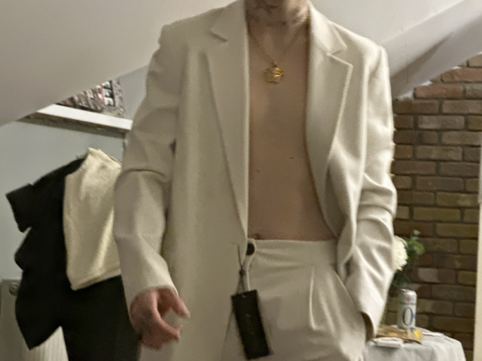 A person stands shirtless, wearing an open, light-colored blazer and matching pants. A gold necklace is visible. A tag hangs from the blazer's button. In the background, there's a brick wall, a table with a can and flowers, and clothing hanging on a rack. The scene is casually staged and appears to be indoors.