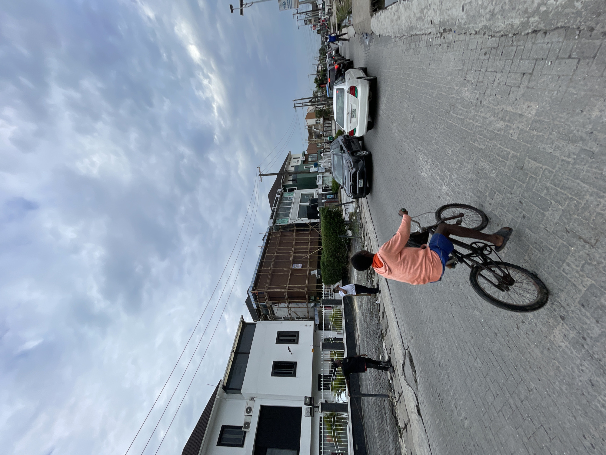 A wide-angle, eye-level shot captures a street scene in Moba, Nigeria, under a cloudy sky. In the foreground, a young boy is riding a bicycle towards the right of the frame. He is wearing an orange hoodie and blue shorts. The road is paved with grey cobblestones.
Along the left side of the street, a white building with black-trimmed windows and air conditioning units stands behind a white metal fence. Next to it, another building is under construction, covered in scaffolding made of bamboo. Two men are standing on the sidewalk near the fence; one in a black t-shirt and jeans, gesturing with his left arm, and another in a white shirt and dark pants, holding his phone to his ear.
The street is lined with more buildings, some appearing to be residential and others commercial. Several cars are visible on the road, including a dark SUV and a white sedan in the middle ground, with more traffic extending into the distance. A street lamp with signs is visible on the right side of the road, and a drainage ditch runs along the edge. The overall impression is of an active urban environment with ongoing development.