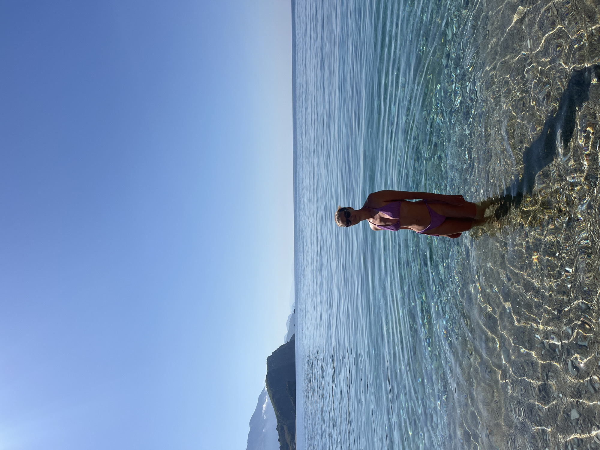 The image depicts a woman standing in shallow, clear water with a sandy or pebbly bottom. She is wearing a purple bikini and sunglasses. The water is calm, reflecting sunlight. Behind her is a vast expanse of blue sky and calm water meeting at the horizon. The location is likely coastal, with a mountain visible in the distance. It could be Budva, Montenegro.