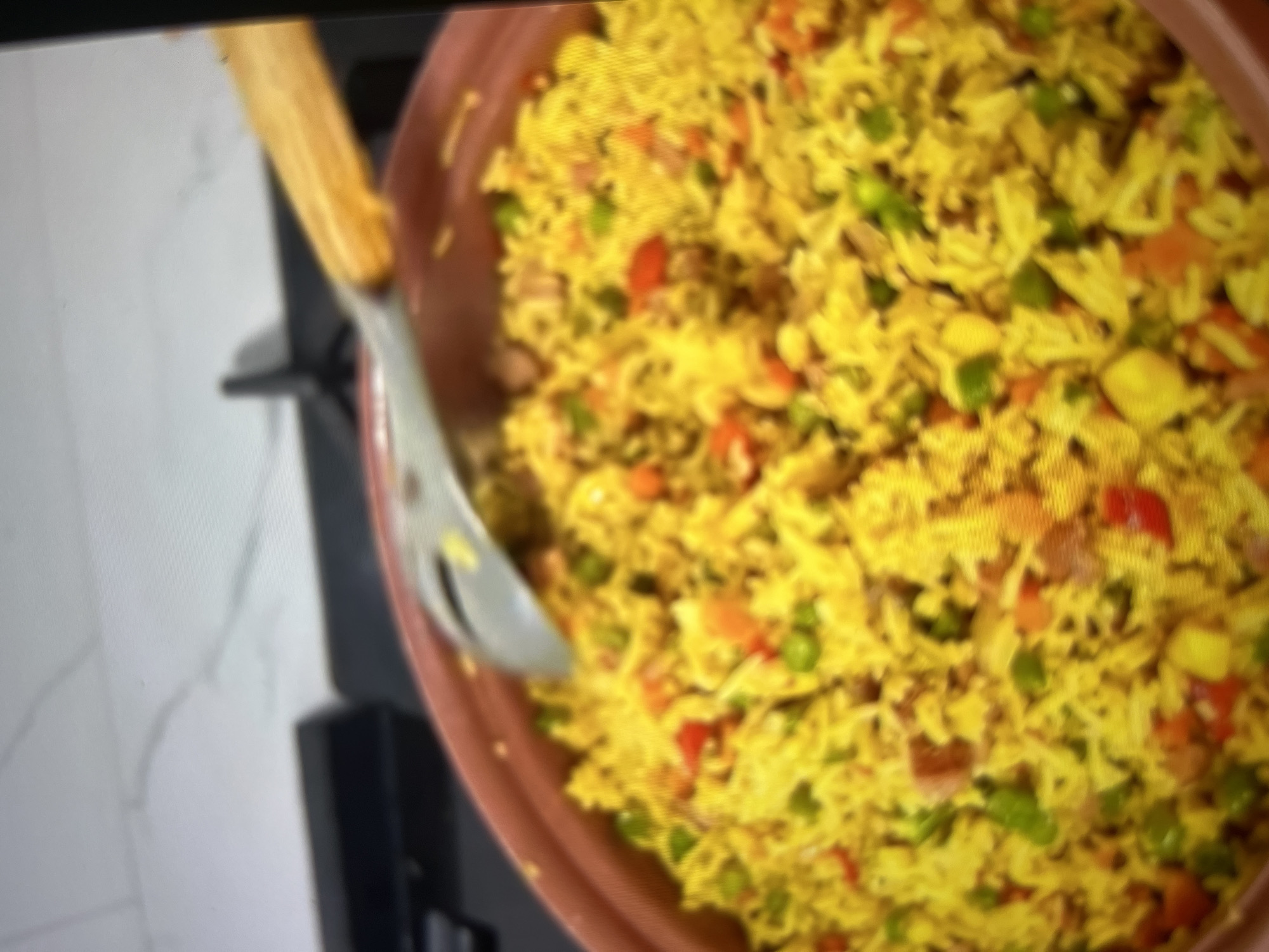 The image shows a close-up of a serving of yellow rice with mixed vegetables, including peas and diced red peppers, in a brown earthenware dish. A wooden-handled serving spoon is partially submerged in the rice. The dish is placed on a black stovetop, and in the background, a white countertop with subtle veining is visible. The scene appears to be a kitchen environment. There is no indication of the city of Abuja, Nigeria, in the image itself.