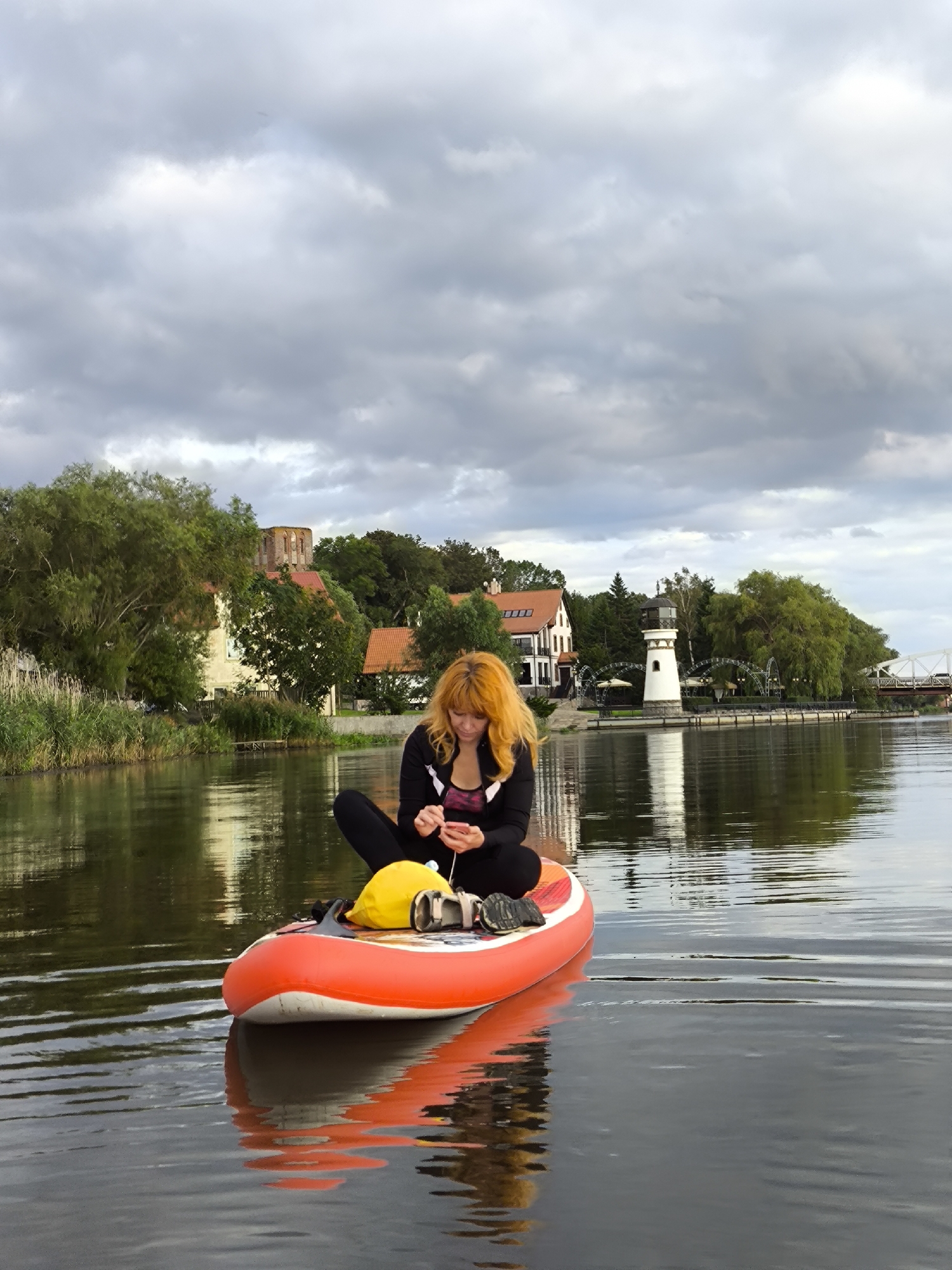 A young woman with bright orange hair, wearing a black jacket over a pink and black sports top and black leggings, sits on an orange paddleboard in calm water. She is holding a smartphone in her hands, seemingly looking at it. A yellow waterproof bag and a pair of black shoes are also on the paddleboard. The paddleboard is positioned in the foreground, reflecting in the water. In the background, across the water, are several buildings, including a white lighthouse, a house with red-tiled roofs, and what appears to be a historical ruin with a brick tower. Lush green trees and foliage line the shore. The sky is overcast with dramatic, grey clouds. The scene is set in Ushakovo, Russia.