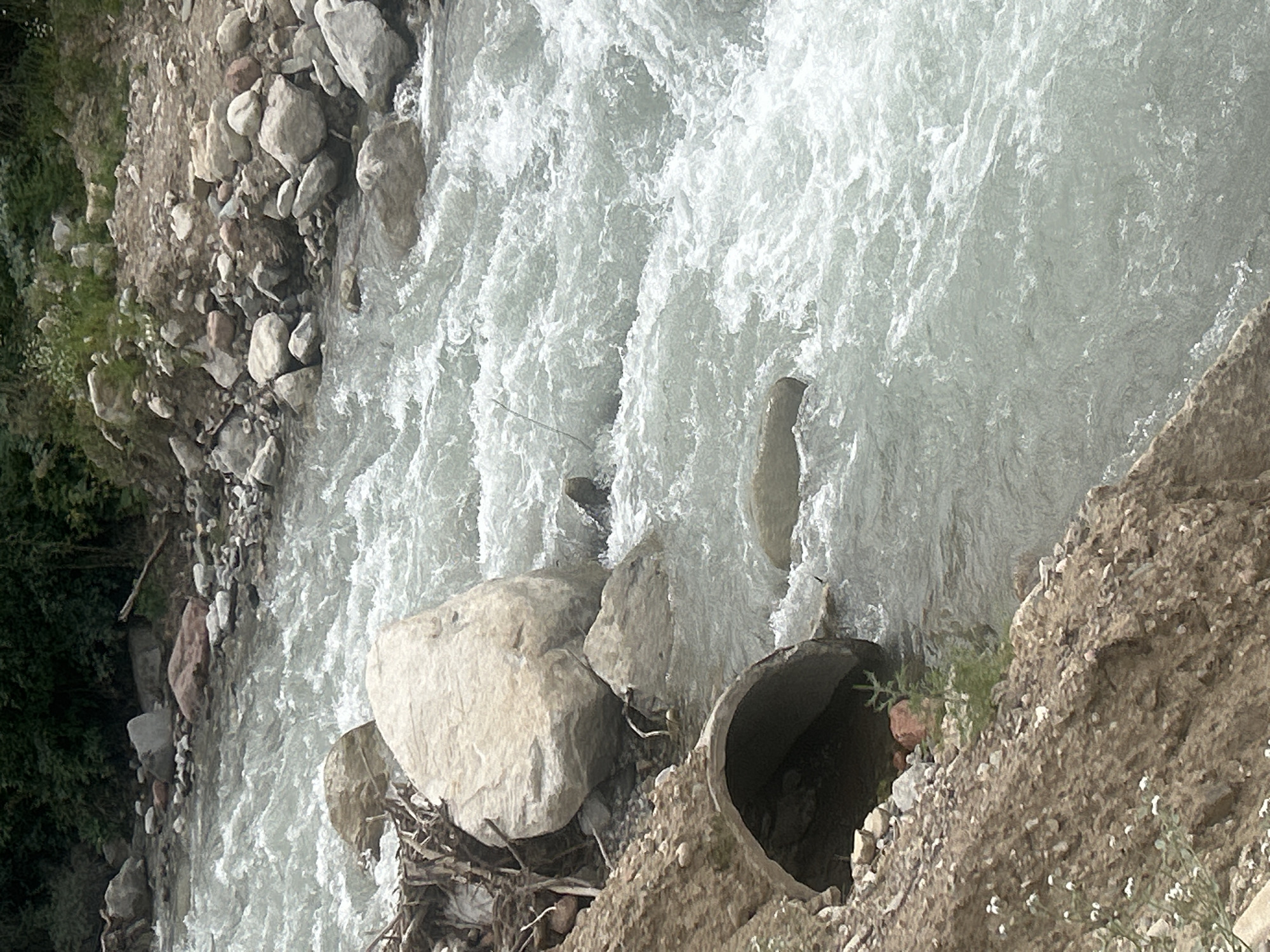 The image shows a turbulent, fast-flowing river with white water rapids. The riverbanks are lined with rocks of various sizes and some dirt. In the foreground, a section of a concrete culvert is visible, partially submerged in the water. Debris, including branches and sticks, is caught near the bank and around some larger rocks. The background shows more rocks and vegetation, with a dense line of trees forming a dark backdrop. The scene appears to be a natural or semi-natural environment within or near the city of Koklaisai, Kazakhstan. There are no people or animals visible in the image.