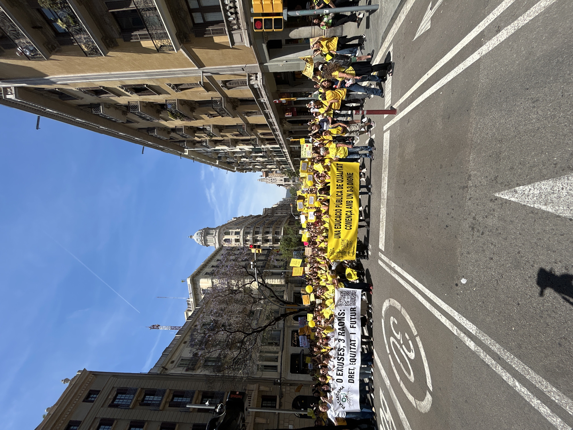 A street-level photograph shows a large group of people marching down a street in Barcelona, Spain. They are mostly wearing yellow shirts and carrying signs and banners, including one that reads "UNA EDUCACIÓ PÚBLICA DE QUALITAT COMENÇA AMB UN 0-3 DIGNE" (A quality public education starts with a 0-3 dignity) and another that reads "0 EXCUSES, 3 RAONS: DRET, EQUITAT I FUTUR" (0 Excuses, 3 Reasons: Right, Equity and Future). The street is lined with buildings, and the sky is blue. The street is marked with a white "30" inside a circle. A traffic light is visible, showing a red light. The "Betevé" logo can be seen near the edge of the first banner.