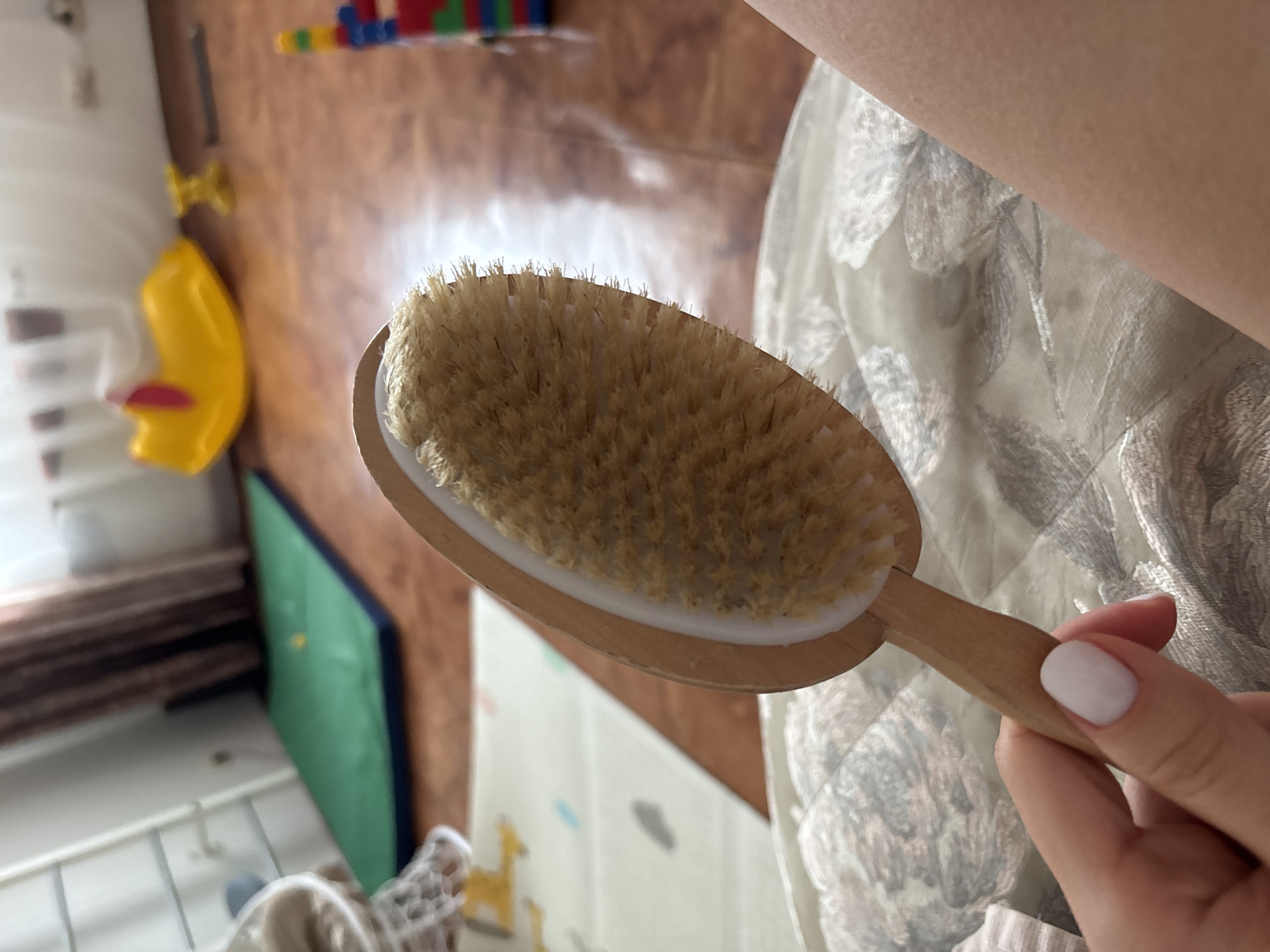 A close-up, high-angle shot captures a person's hand holding a wooden dry brush with natural, light-brown bristles. The hand has light-toned skin and a white-painted thumbnail. The brush features an oval head with a white plastic or silicone rim and a short, light-colored wooden handle.
The immediate background to the right and below the hand shows a patterned fabric, possibly a blanket, curtain, or clothing, with a white and gray floral or abstract design.
The rest of the background is blurred, indicating an indoor setting, likely a domestic space such as a child's room or a play area. Visible elements in the background include a bright yellow plastic toy, possibly a slide, resting on a brown wooden floor or furniture. Above this, colorful building blocks (red, blue, green, yellow) are arranged on a surface. A green mat or cushion is seen vertically on the left side of the blurred background, and part of a light-colored playmat with a faint giraffe outline is visible further down. The lighting suggests daytime indoors. No specific activity beyond holding the brush is discernible. No text is visible in the image.