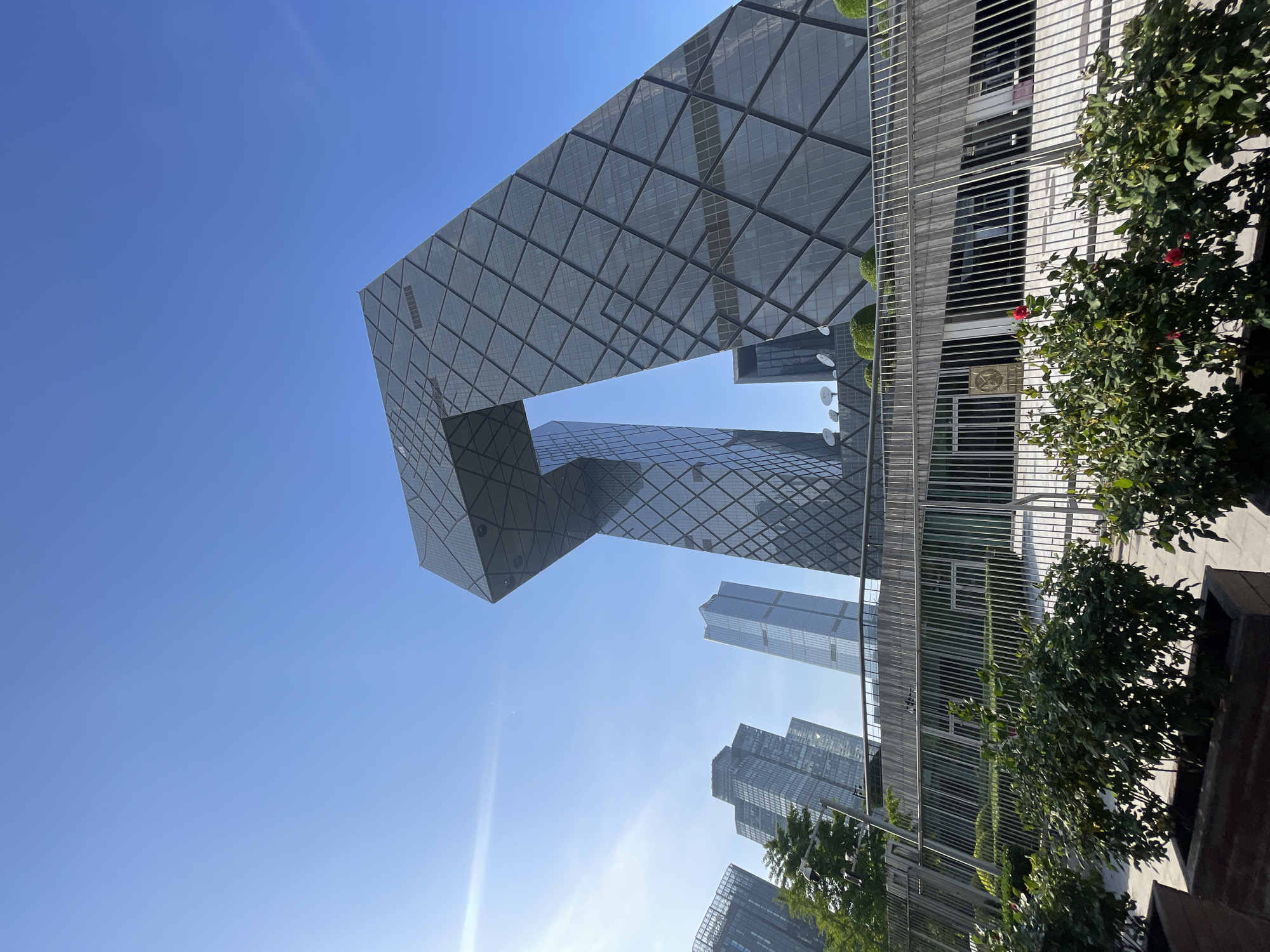 The image shows a daytime view of the China Central Television (CCTV) Headquarters in the Chaoyang District of Beijing, China. The unique loop-like structure of the building is the primary subject. Other skyscrapers are visible in the background. A metal fence and greenery are present in the foreground. The sky is clear and blue.