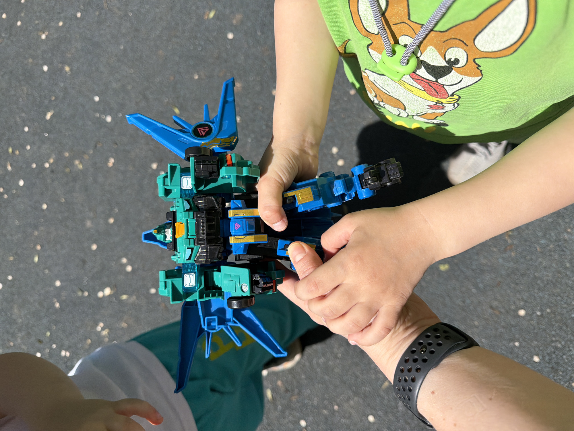 The image shows a close-up, outdoor shot of two children holding a blue and green toy robot. One child's hands are on the robot's legs, while the other child's hands are on its body. A third child is partially visible in the background to the left, wearing a white shirt and teal shorts. The ground appears to be asphalt with scattered leaves. The lighting suggests it is daytime, with a strong shadow cast by one of the children. The scene does not contain any information that would indicate its location in Khabarovsk, Russia.