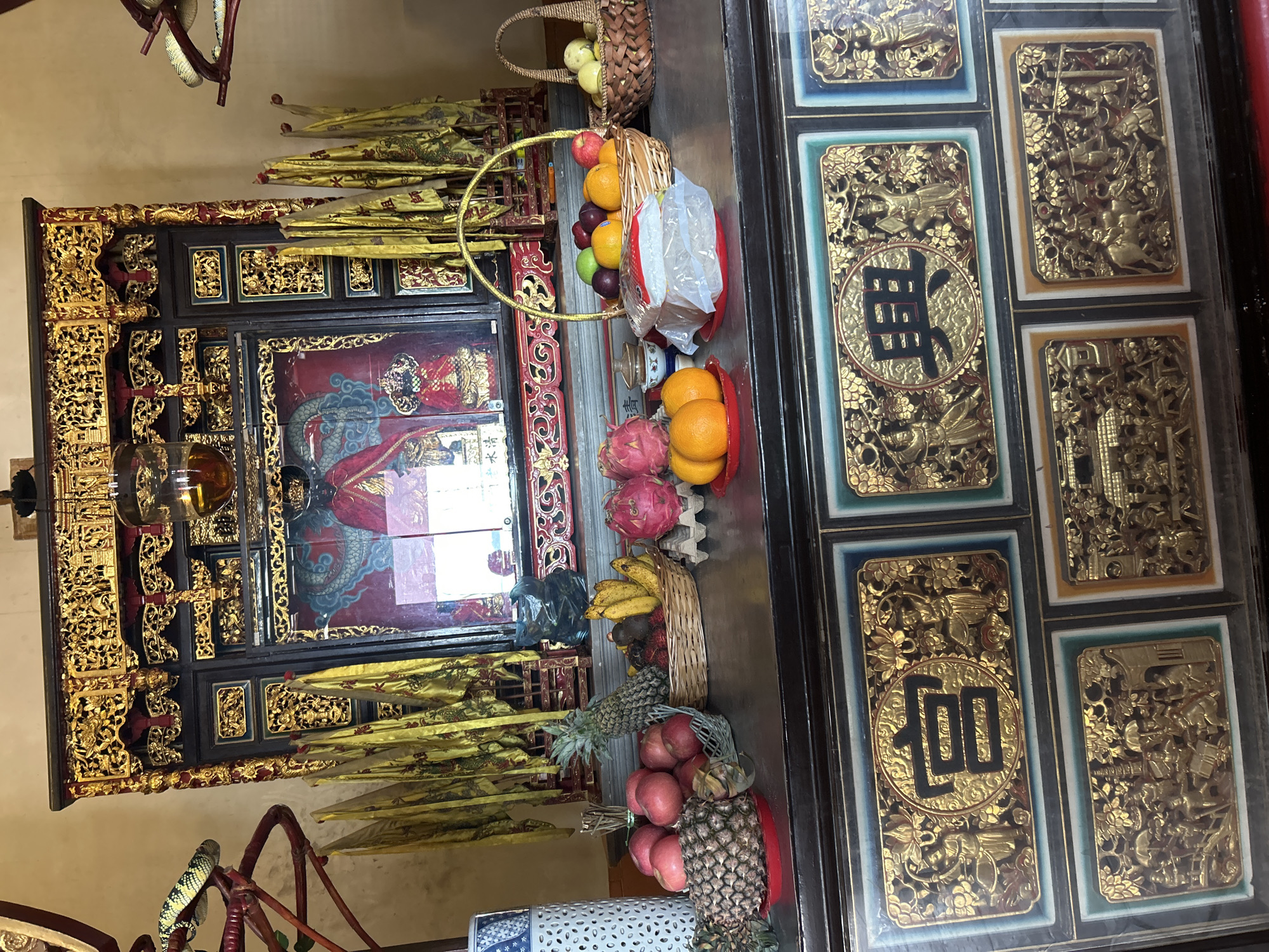 This image depicts an altar dedicated to a deity, likely within a Chinese temple in George Town, Malaysia, given the intricate carvings and offerings. The altar is adorned with golden relief carvings on a dark wood cabinet. These carvings feature figures in traditional attire, mythical creatures like dragons, and scenes of daily life or religious ceremonies.
Dominating the altar is a large, ornate frame surrounding a painting of a deity dressed in red robes. Yellow banners with inscriptions hang on either side. In front of the altar, a variety of offerings are arranged on the cabinet. These include several pineapples, a bunch of bananas, rambutan, apples, dragon fruits, and oranges. Some fruits are placed in woven baskets, while others are on red plates. A small stack of what appears to be white rice, wrapped in plastic, sits on a red plate.
The overall impression is one of reverence and tradition, highlighting the cultural and religious practices prevalent in George Town. The lighting suggests an indoor setting, possibly a dimly lit temple interior.