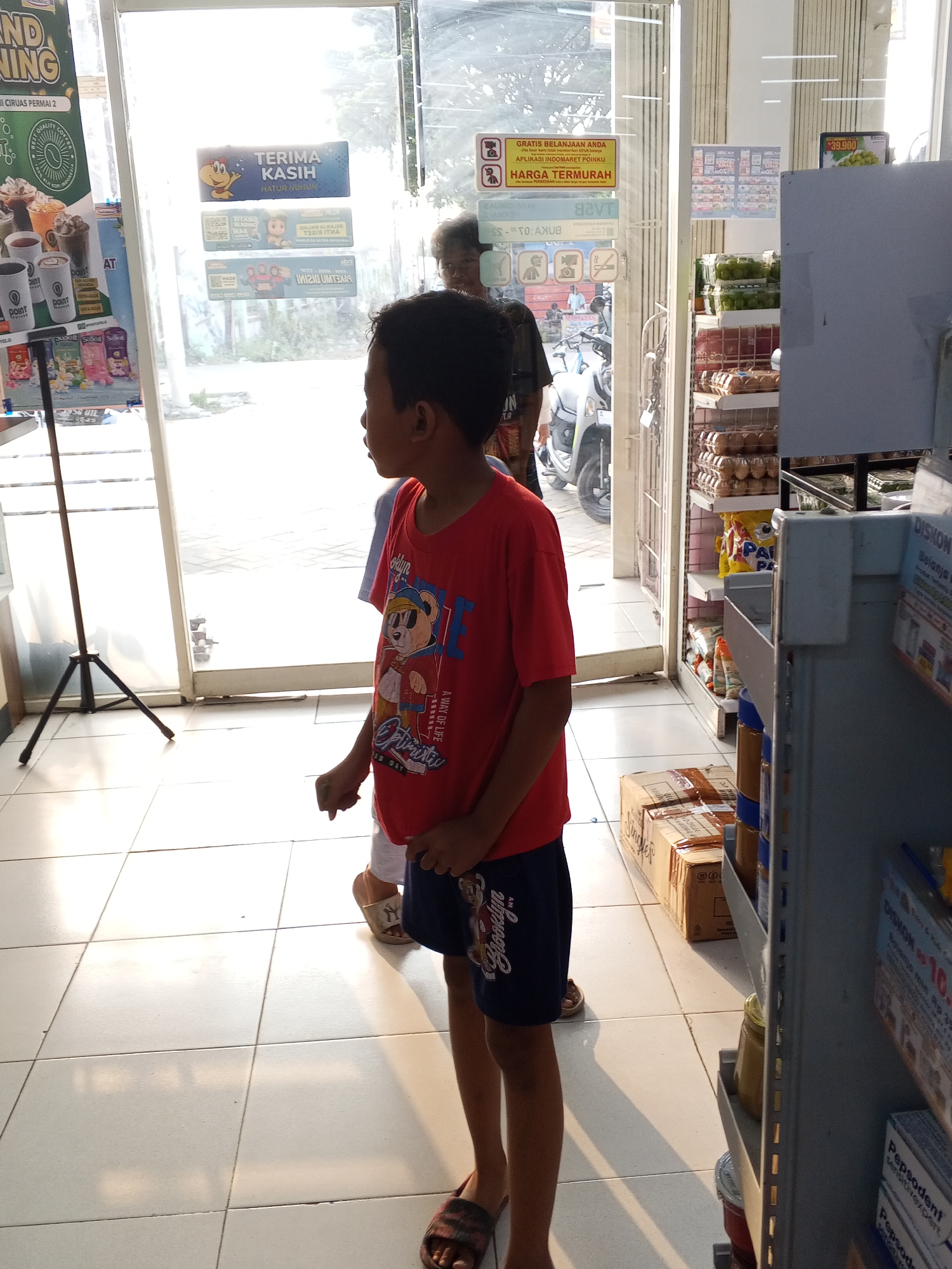 A young boy wearing a red t-shirt with a graphic of a cartoon bear and dark blue shorts is standing near the entrance of a convenience store. He is looking to the right. Behind him, another person, partially visible, is also looking towards the store. The store entrance features glass doors and windows, through which sunlight is streaming. Several signs are visible on the doors, including one that says "TERIMA KASIH" (Thank You) and another with "GRATIS BELANJA ANDA" (Your Shopping is Free) and "HARGA TERMURAH" (Cheapest Price). Inside the store, shelves are stocked with various products, including eggs, packaged food items, and cleaning supplies. A motorcycle is parked outside the entrance. The floor is tiled with square white tiles. The overall scene depicts a typical daytime scene at a retail establishment in Ranjeng, Indonesia.
