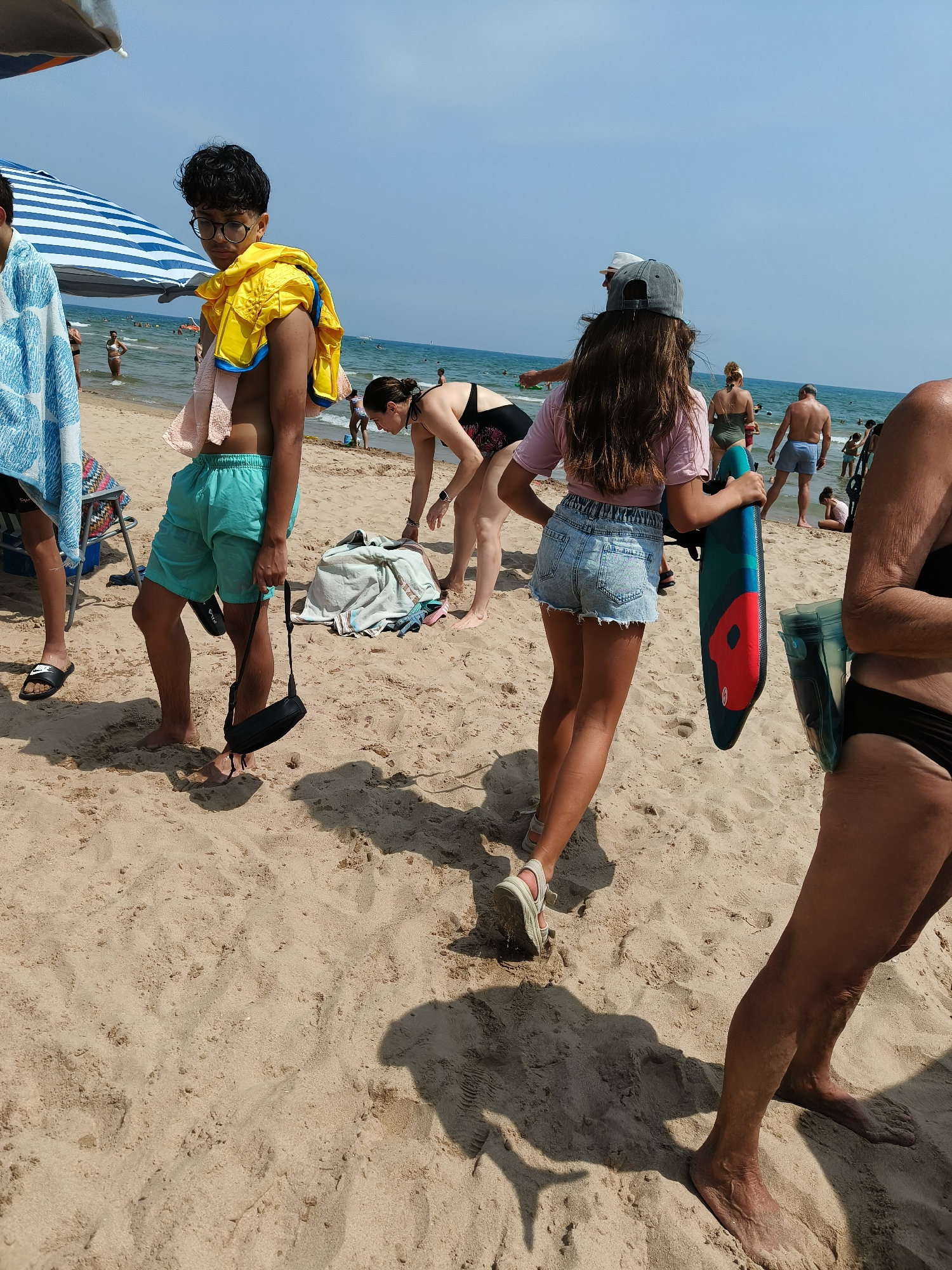 The image depicts a sunny day at the Gandia beach in Spain. Several people are present on the sandy shore. In the foreground, a young person wearing turquoise shorts and a yellow life vest draped over their shoulder is standing, holding a black bag. To their right, another person is holding a colorful boogie board and wearing denim shorts. Further in the background, individuals are seen walking or relaxing near the water. The sea is calm and extends to the horizon under a clear blue sky. Beach umbrellas and towels are scattered on the sand, indicating a typical beach scene with recreational activities. The sand is light brown and appears dry, with footprints and shadows cast by the people and objects.