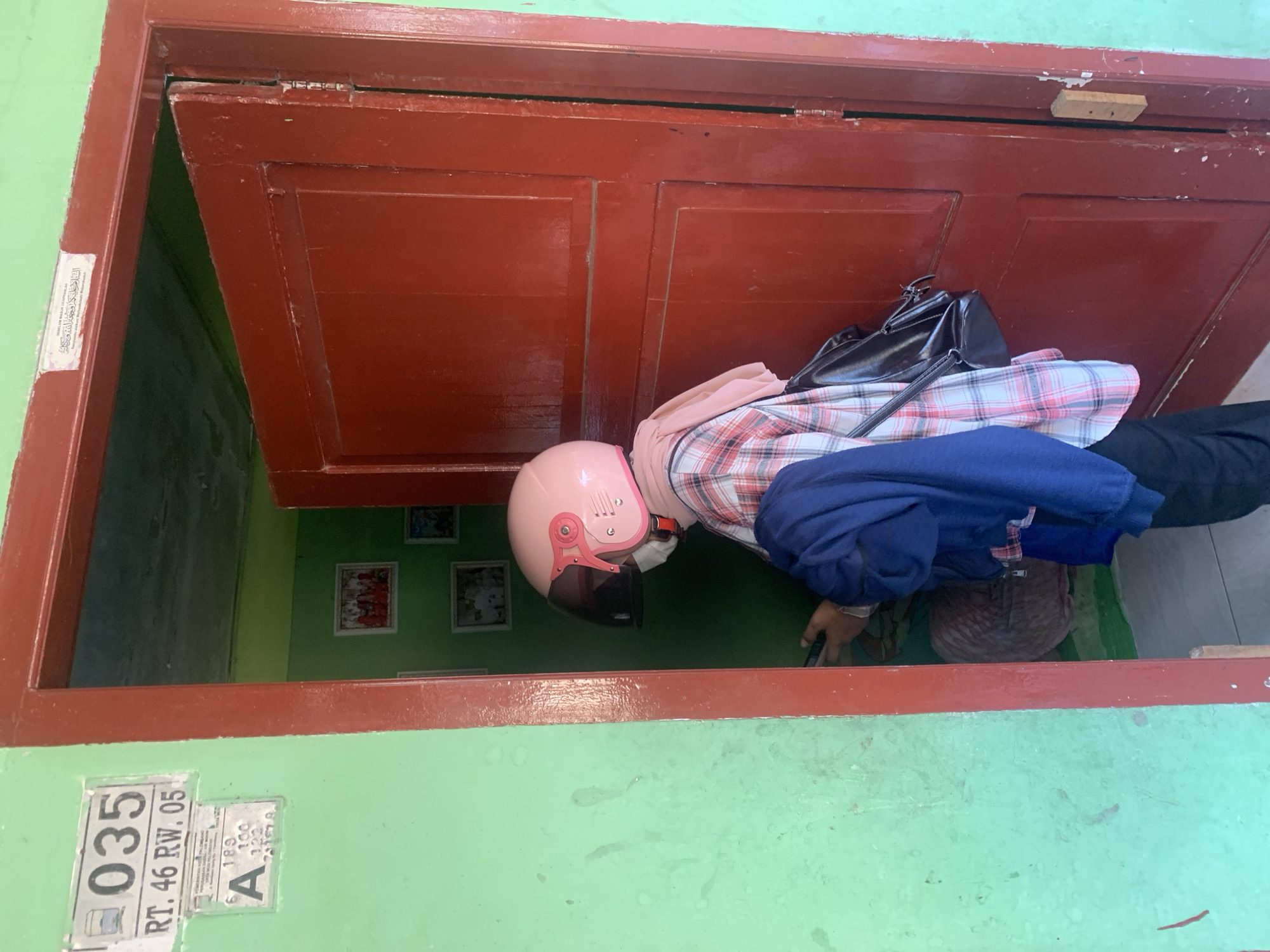 A person wearing a pink helmet and a plaid shirt is standing in a doorway. The person is holding a phone and a dark blue jacket. A black backpack is slung over their shoulder. The doorway leads into a room with green walls and several framed pictures. A sign on the green wall outside the doorway reads "035 RT. 46 RW. 05". The door itself is red and appears to be made of wood. The scene is set in Palembang, Indonesia, as indicated by the address on the sign.