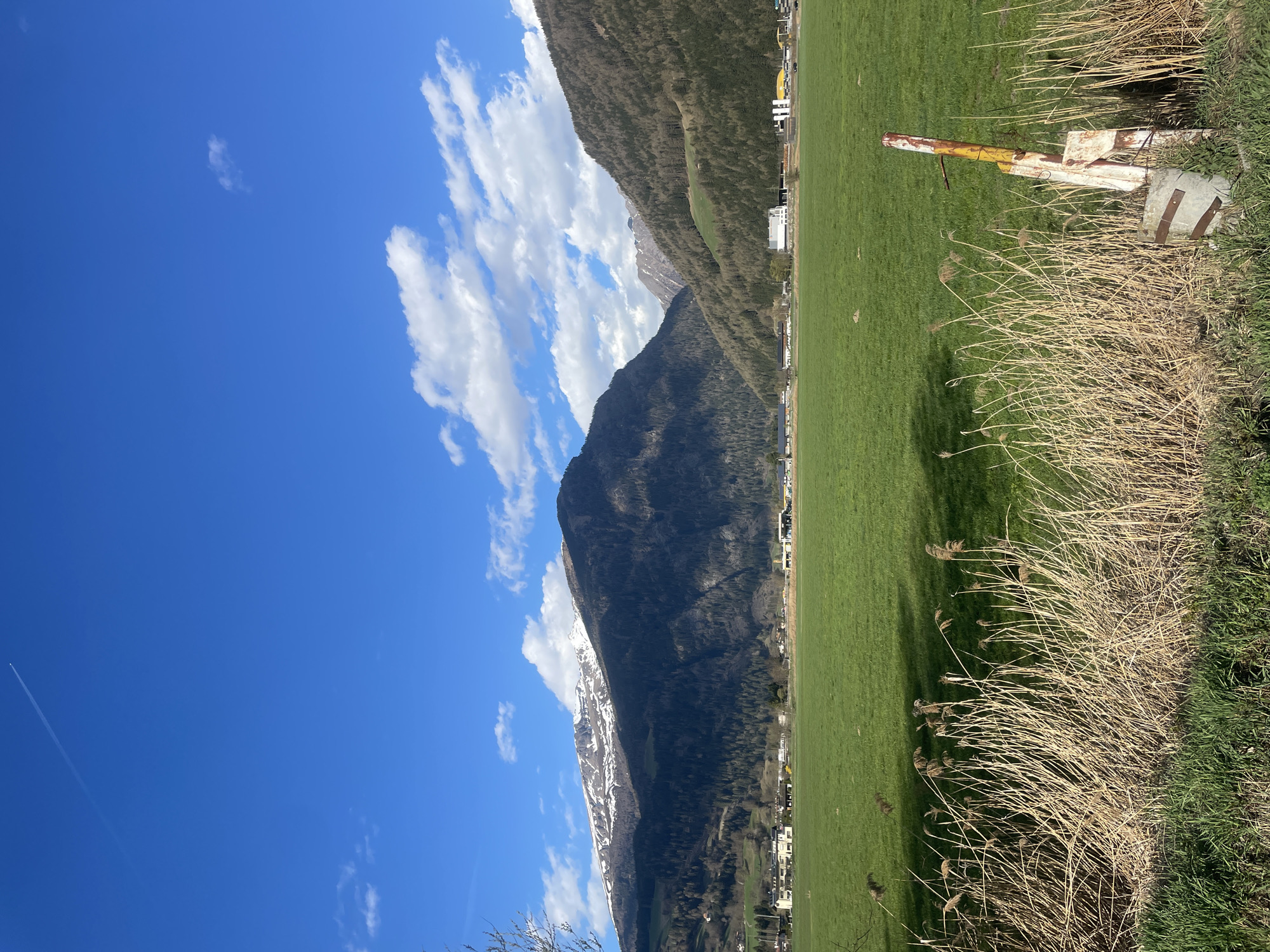 The image depicts a landscape in Sterzing-Vipiteno, Italy, featuring a clear blue sky with scattered cumulus clouds. A mountain range, partially covered in snow, dominates the left side of the frame, while a grassy field occupies the right. Along the edge of the field, there's a patch of dry, brown grass and some green vegetation. In the foreground, near the grass, there's a weathered, yellow and white metal structure. A contrail from a plane can be seen in the upper left quadrant of the image. The environment suggests a rural or suburban area.