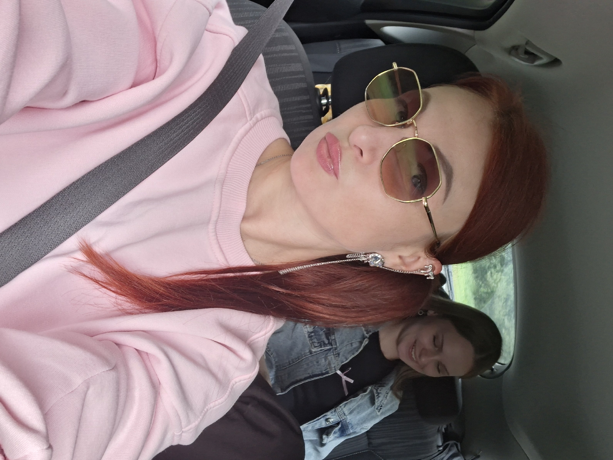 Two women are in the back seat of a car, presumably traveling. The woman in the foreground, with red hair and sunglasses, is taking a selfie. She is wearing a pink sweatshirt and a seatbelt. She has a dangling earring with a chain. The woman in the background, with light brown hair, is looking down and smiling. She is wearing a denim jacket over a black t-shirt. The scene appears to be during the day, with greenery visible outside the car window. The location is identified as Yunkavichy rural council, Belarus.
