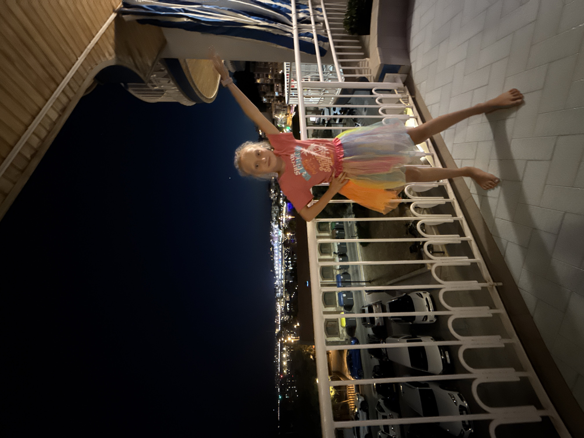 A young girl with blonde hair, wearing a pink t-shirt and a multi-colored tutu, stands on a tiled balcony at night, her right arm extended upwards and her left hand on her hip. Behind her, a distant city skyline is illuminated with numerous lights, suggesting the location is Blanes, Spain. A white railing separates the balcony from a street below where several cars are parked. The sky is dark, and a portion of the balcony's wooden ceiling and a striped awning are visible. The girl is barefoot.