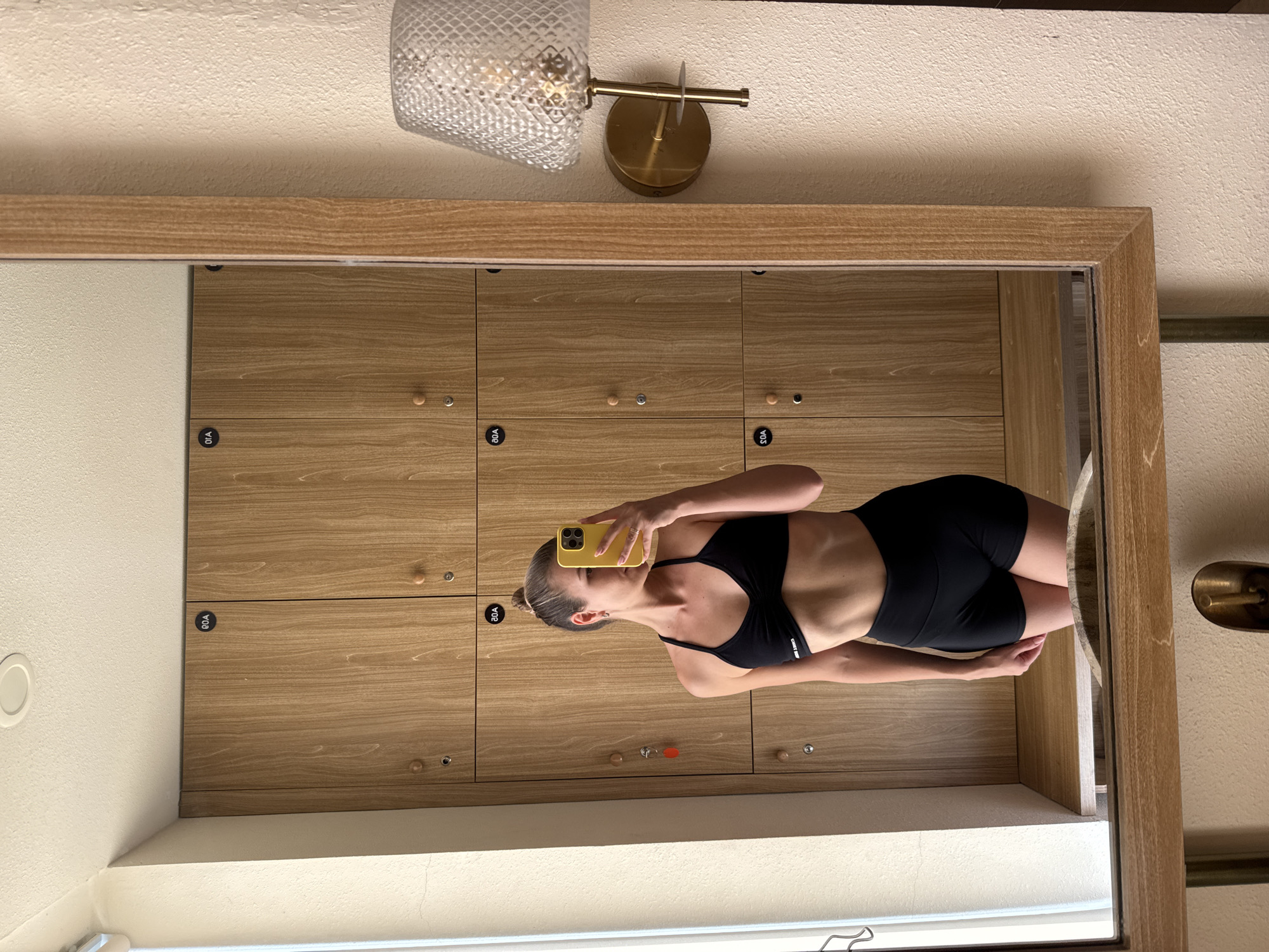 A person is taking a selfie in front of a mirror. The mirror is framed with light-colored wood and reflects a series of wooden lockers. The lockers have small, round knobs and small, black number plates. Above the mirror, there is a decorative light fixture with a glass shade and a brass-colored base. The surrounding walls are painted in a neutral, light tone.
