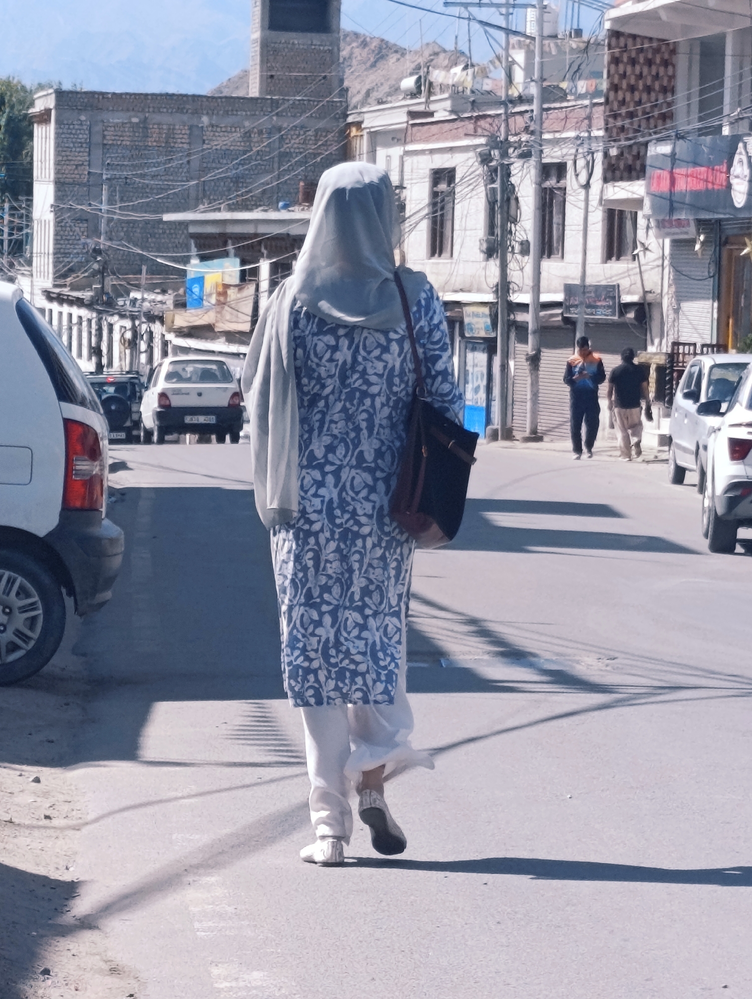 The image depicts a street scene in Leh, India, under a bright, sunny sky. A woman, seen from the back, is walking away from the camera, wearing a light grey hijab and a blue and white patterned dress with white pants. She carries a dark brown shoulder bag. To her left, a white car is partially visible. Further down the street, several other vehicles are parked or in motion, including a white Maruti car with a license plate, and other cars further in the background. Two men are walking on the right side of the street. Buildings line the street, some with a rustic, stone construction, and others with more modern facades. Power lines are visible crisscrossing above the street and between buildings. The overall environment suggests a bustling, day-time activity on a street in a town or city.