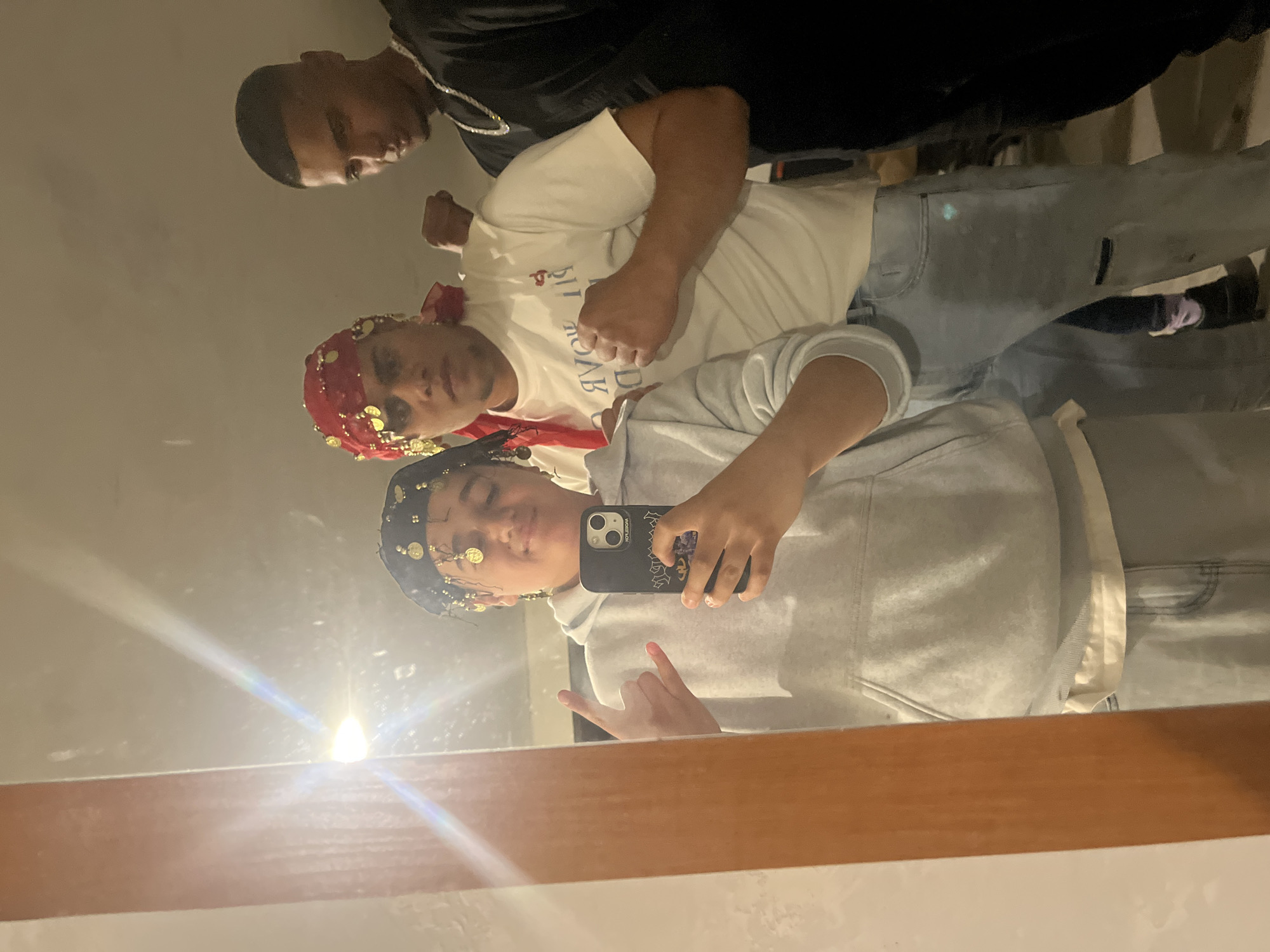Three individuals are visible in a mirror selfie. The foreground figure wears a blue head covering adorned with gold discs, a grey hooded sweatshirt, and is holding a smartphone in their right hand while making a hand gesture with their left. Behind them is another person wearing a red head covering with gold embellishments and a white t-shirt. A third person, partially visible in the upper left, is wearing a black jacket and a chain. The scene is indoors, reflected in a mirror with a wood frame. The location is not evident from the image.
