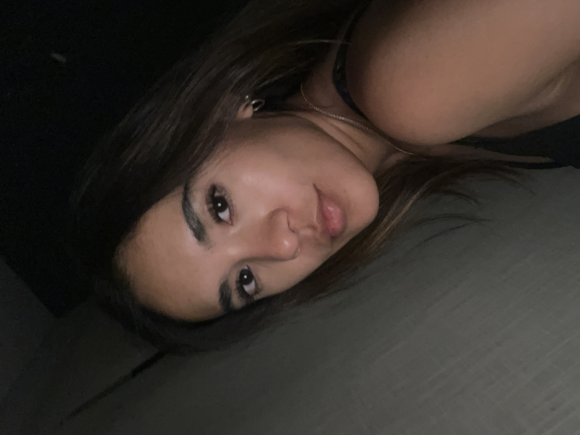 A young woman lies on a gray surface, possibly fabric or flooring. Her head is tilted towards the camera, and she looks directly at the viewer. She has dark brown hair, brown eyes, and fair skin. She is wearing a dark-colored sleeveless top. A small hoop earring is visible in her left ear, and a thin necklace is around her neck. The background is dark and indistinct. There is no indication of the location being San Bernardino Tlaxcalancingo, Mexico.