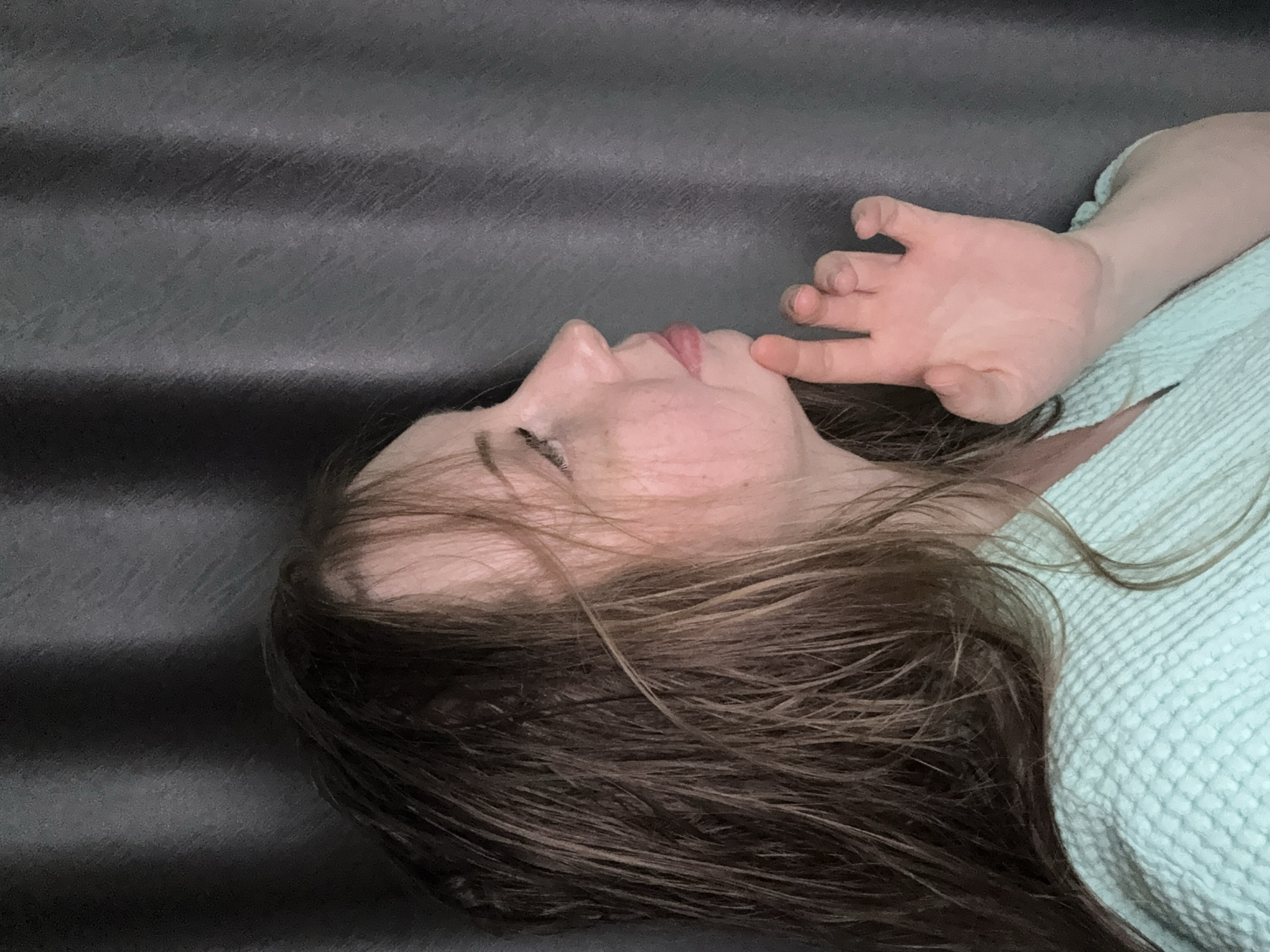 A person is shown in a mid-angle shot, lying on their back with their head turned to the left, revealing their profile. Their eyes are closed or partially closed, and their lips are slightly parted. Long, brown hair, appearing somewhat disheveled and wavy, is spread out around their head.
The person is wearing a light blue garment with a noticeable textured, possibly waffle-knit, pattern. Their right hand is raised, resting gently near their face, with fingers slightly curled against their chin or cheek. The posture and closed eyes suggest a state of rest, relaxation, or sleep.
The background is a dark, solid-colored surface, likely grey or black, featuring horizontal ridges or folds. This suggests an indoor setting, possibly a bed or a couch. The lighting is soft and diffused, contributing to a calm and peaceful atmosphere. No text is visible in the image.
