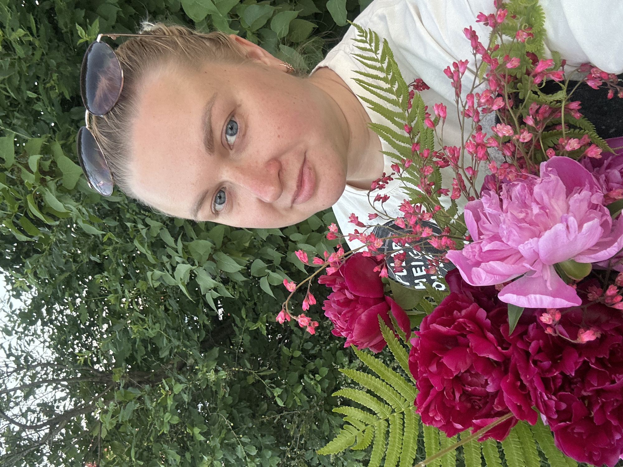 The image depicts a woman holding a floral bouquet in an outdoor setting, situated in the city of Chaikovsky, Russia. The woman is presented in a close-up, chest-up shot, looking directly at the viewer with a neutral to slightly smiling expression. She has light-toned skin, blue eyes, and light-colored hair pulled back from her face. Sunglasses with dark, oval-shaped lenses and gold-colored frames are perched on top of her head. A small gold-toned hoop earring is visible in her right ear. She wears a white crew-neck t-shirt, part of which features a dark graphic print, partially obscured by the flowers, reading "FLOP HEIG..." or similar.
She holds a large, dense bouquet of flowers and foliage in front of her torso, centered slightly to her left. The bouquet primarily consists of several large, vibrant deep magenta/fuchsia-colored peonies and at least one lighter pink peony, alongside multiple bright green fern fronds. Smaller, delicate pink bell-shaped flowers on slender, branching stems are also integrated into the arrangement.
The background is dominated by lush, dense green foliage, appearing as a mixture of trees and bushes, suggesting a garden or natural outdoor environment. Glimpses of tree trunks are visible through the leaves on the left side. The lighting is soft and even, suggesting an overcast day or shaded conditions. A sliver of light-colored sky is visible in the upper left corner of the frame.