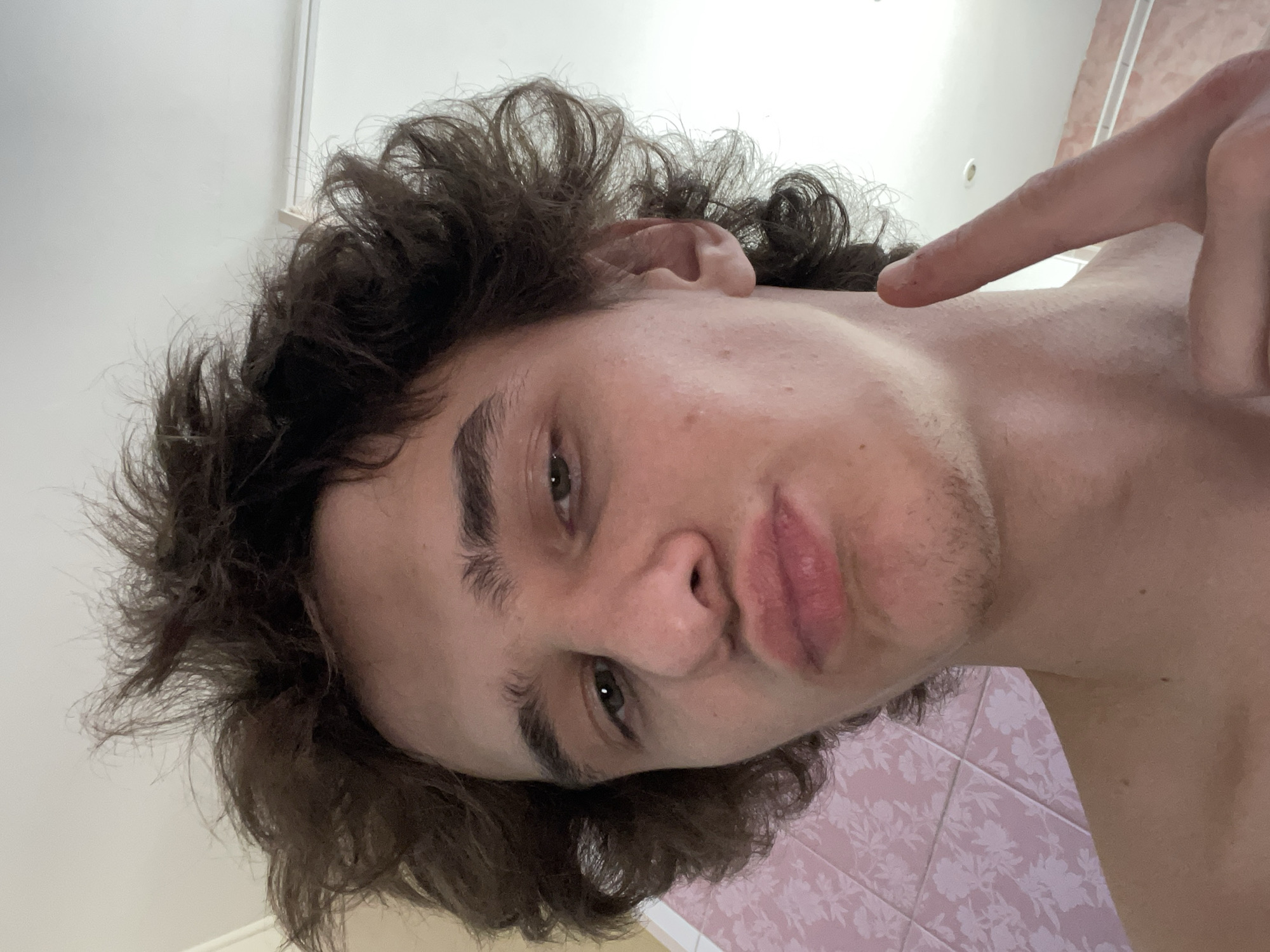 A young man with curly brown hair is pointing a finger at his neck. He has light skin, brown eyebrows, and stubble. His lips are pursed slightly. He appears to be in a bathroom with pink tiled walls and a white ceiling. The location of the image is unknown, and the image contains insufficient information to determine if the subject is in Krasnoyarsk, Russia.