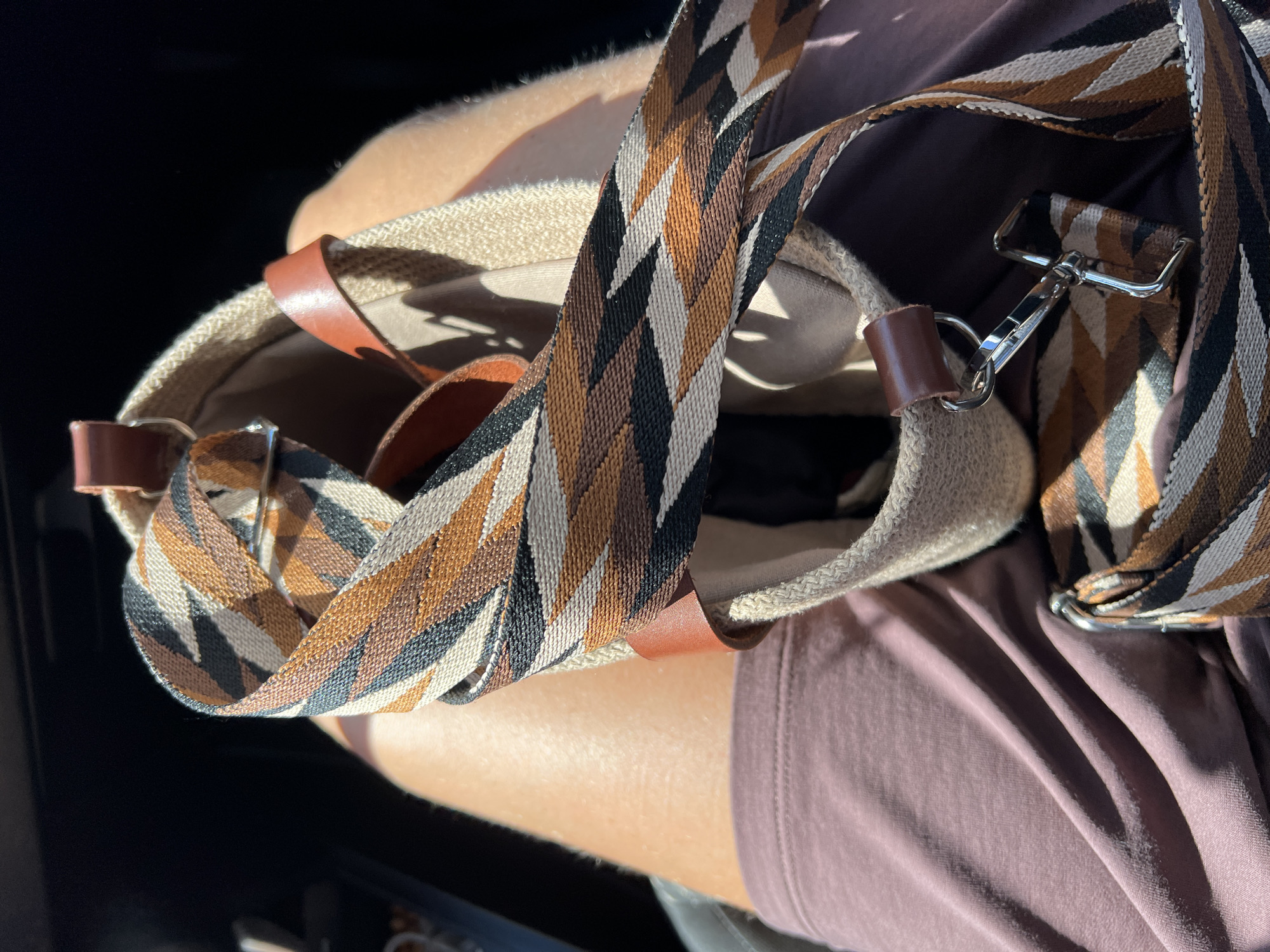 The image shows a close-up, overhead view of a woman's legs and a partially visible canvas tote bag. The woman is wearing a mauve-colored dress or skirt that is wrinkled. The bag has a woven strap with a geometric pattern of brown, black, and tan. A leather strap detail is visible on the bag, as well as a silver metal clip. The bag appears to be sitting on the woman's lap, suggesting she may be in a vehicle or sitting down. The background is dark and out of focus, indicating a low-light environment. The lighting suggests the photograph was taken outdoors during the day, with harsh sunlight casting shadows. There is no discernible information within the image to suggest the location is Salavat, Russia.