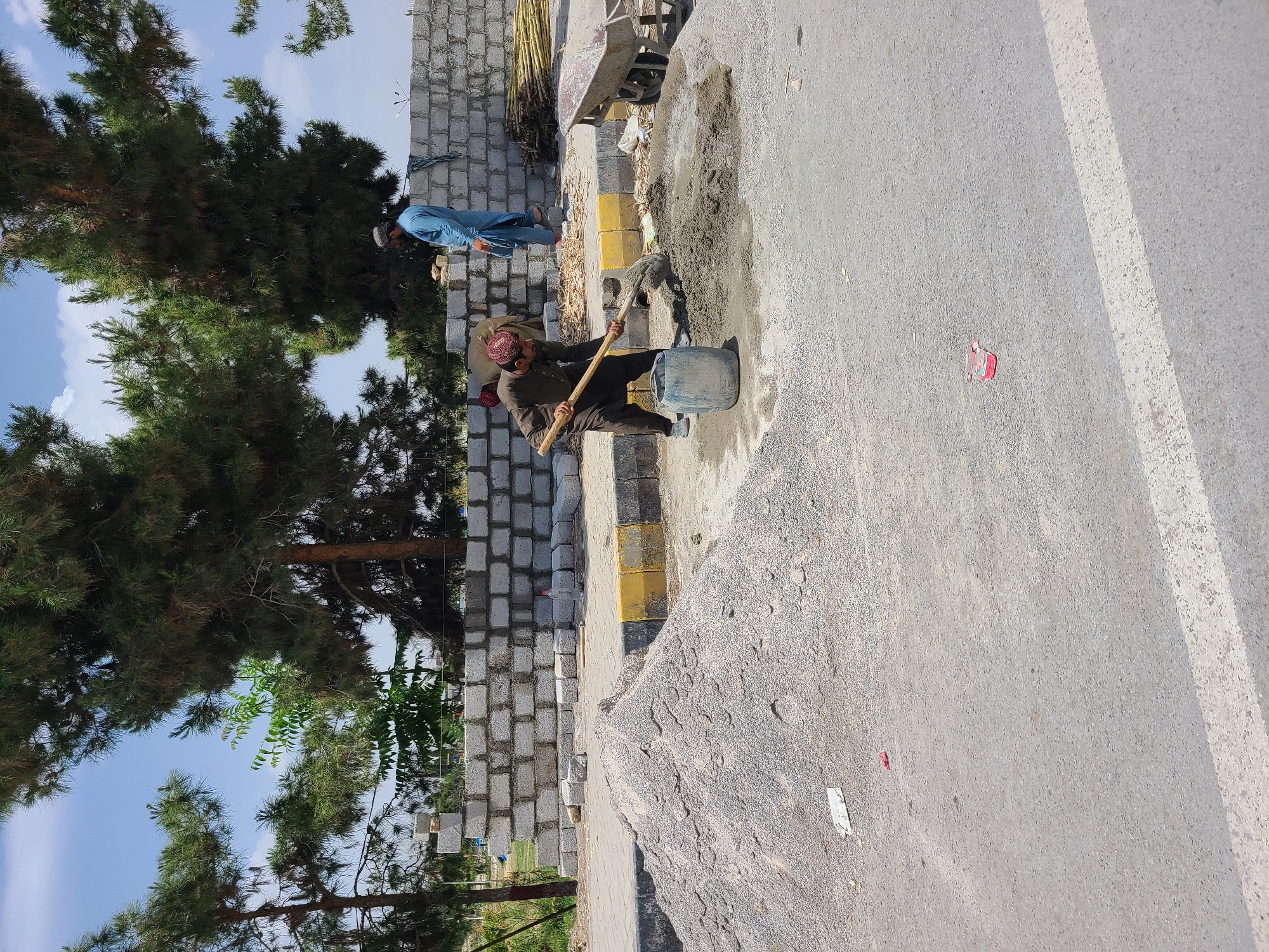 This outdoor image, likely taken in Quetta City Tehsil, Pakistan, depicts a scene of construction or road maintenance under a clear, bright blue sky with some white clouds. In the foreground, a man dressed in a dark traditional outfit and a patterned cap is actively mixing concrete or mortar with a shovel on a dirt and gravel patch. He uses a blue plastic bucket, possibly for water or to hold materials, while a fresh pile of concrete mix is visible on the ground beside him.
Behind this individual, another man in a light blue shalwar kameez and a white cap is partially visible near a newly constructed grey concrete block wall. This wall runs alongside a yellow-painted curb, separating the work area from a paved road. Adjacent to the curb, a substantial pile of gravel or small stones is present, along with a wheelbarrow that holds bundles of what appear to be bamboo or reeds.
The paved road shows a distinct white marking line and has minor debris, including a small red plastic item and a piece of white paper. In the background, large, vibrant green pine trees and other foliage create a natural backdrop. The presence of clear shadows indicates a sunny daytime setting. The overall scene captures diligent manual labor in a typical construction environment. No visible text is legible in the image.