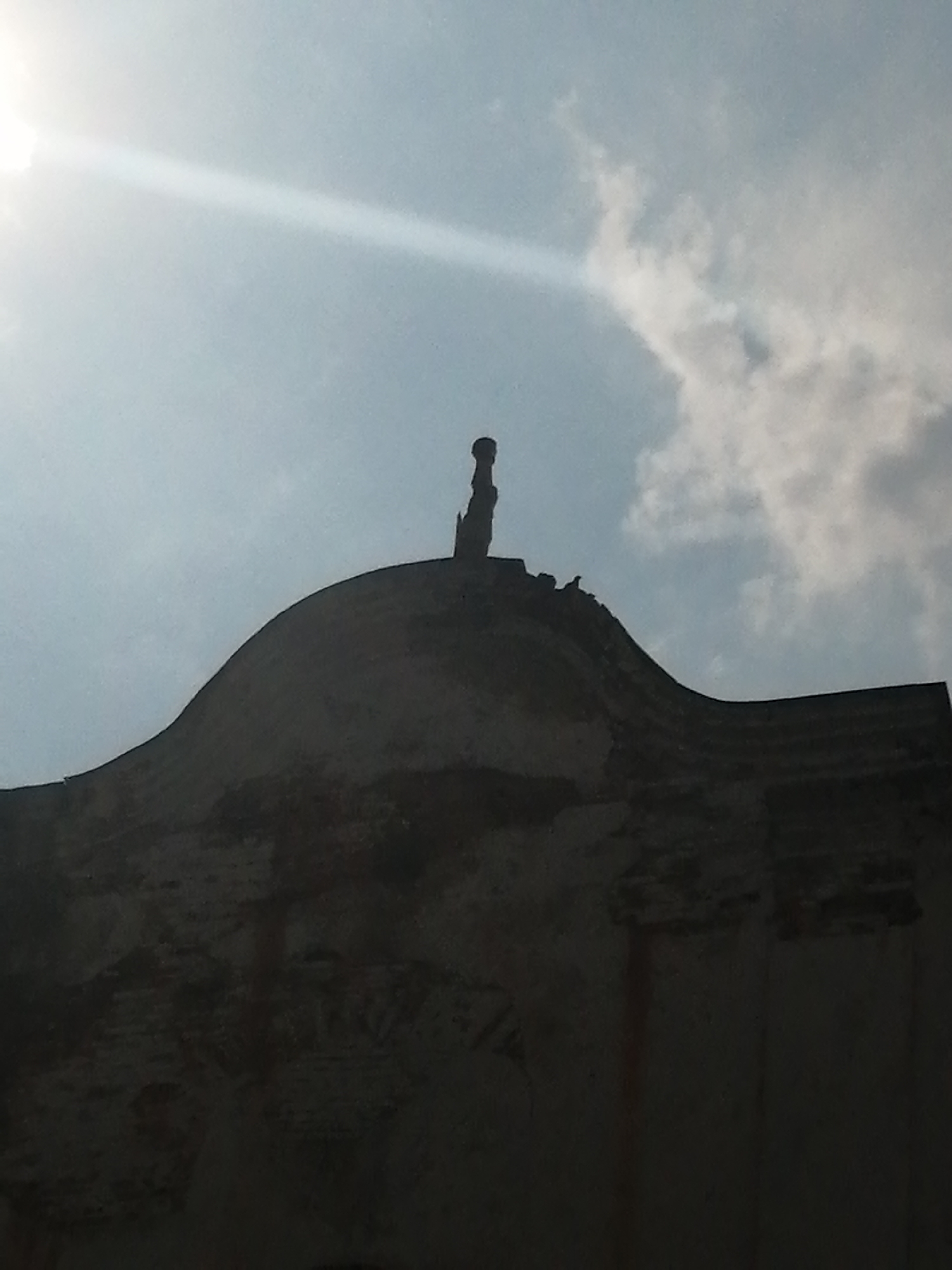 This is a low-angle, silhouetted photograph of the roof and upper facade of a weathered, possibly historic building in Slonim, Belarus. The sky above is a pale blue with scattered clouds and a bright sunray emanating from the upper left corner. Perched on the edge of the roof, silhouetted against the sky, are two birds. One is a small, dark bird, and the other is a slightly larger, dark bird with its wings tucked. At the apex of the curved roof, a small, dark, finial-like object is visible. The building's surface appears to be made of brick, with some areas showing signs of decay and visible texture. The overall impression is one of age and perhaps neglect, with the birds adding a touch of life to the scene.