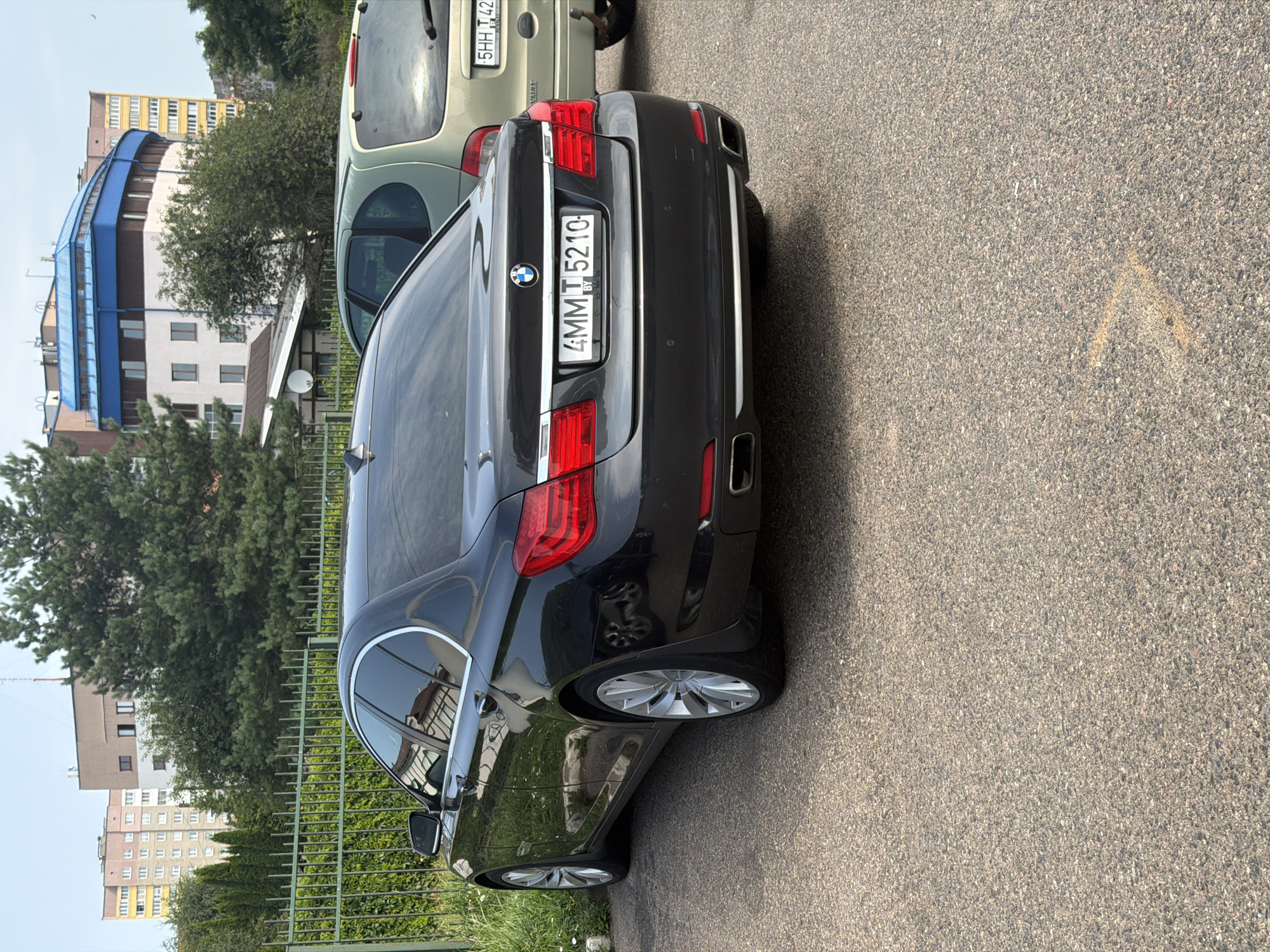In Minsk, Belarus, a black BMW sedan is parked on an asphalt surface. The car's license plate reads "4MMT 5210 BY". Behind the BMW, a light-colored minivan is also parked. The background features buildings and trees, with a green metal fence separating the parking area from the grassy area behind it. The sky is visible above the buildings. The scene appears to be a typical urban environment.