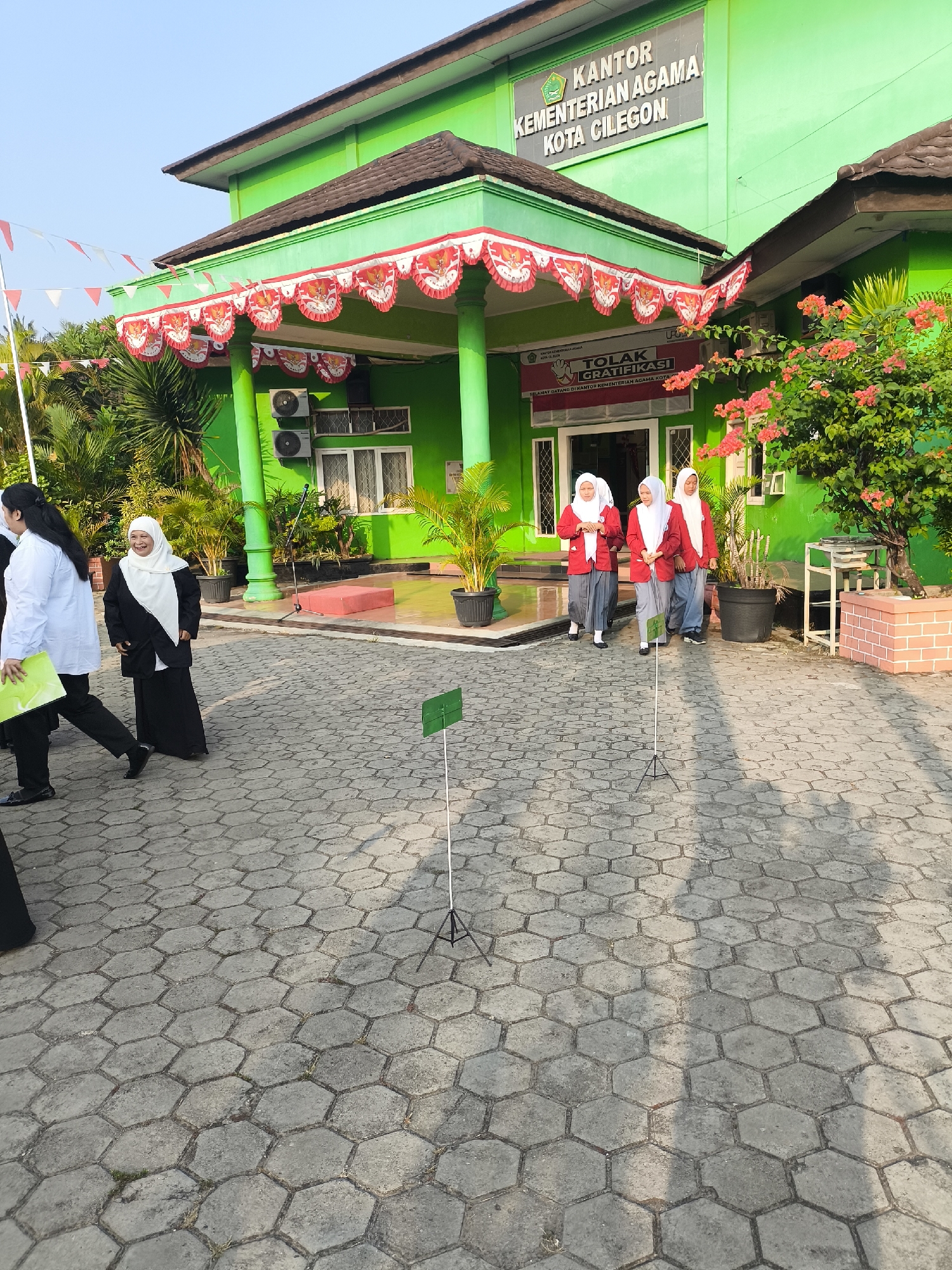 The image shows the front entrance of the KANTOR KEMENTERIAN AGAMA KOTA CILEGON in Cilegon, Indonesia. The building is painted bright green with a red and white decorative banner hanging under the eaves. Several people are visible on the paved courtyard in front of the building. On the left, two women are walking, one in a white shirt and black pants, the other in a black and white outfit, smiling. Three young women in hijabs and red tops are walking towards the entrance. The ground is paved with hexagonal gray stones. Two green signs on white poles are planted in the ground. A tree with pink flowers is on the right side of the image. The sky is clear and blue.
