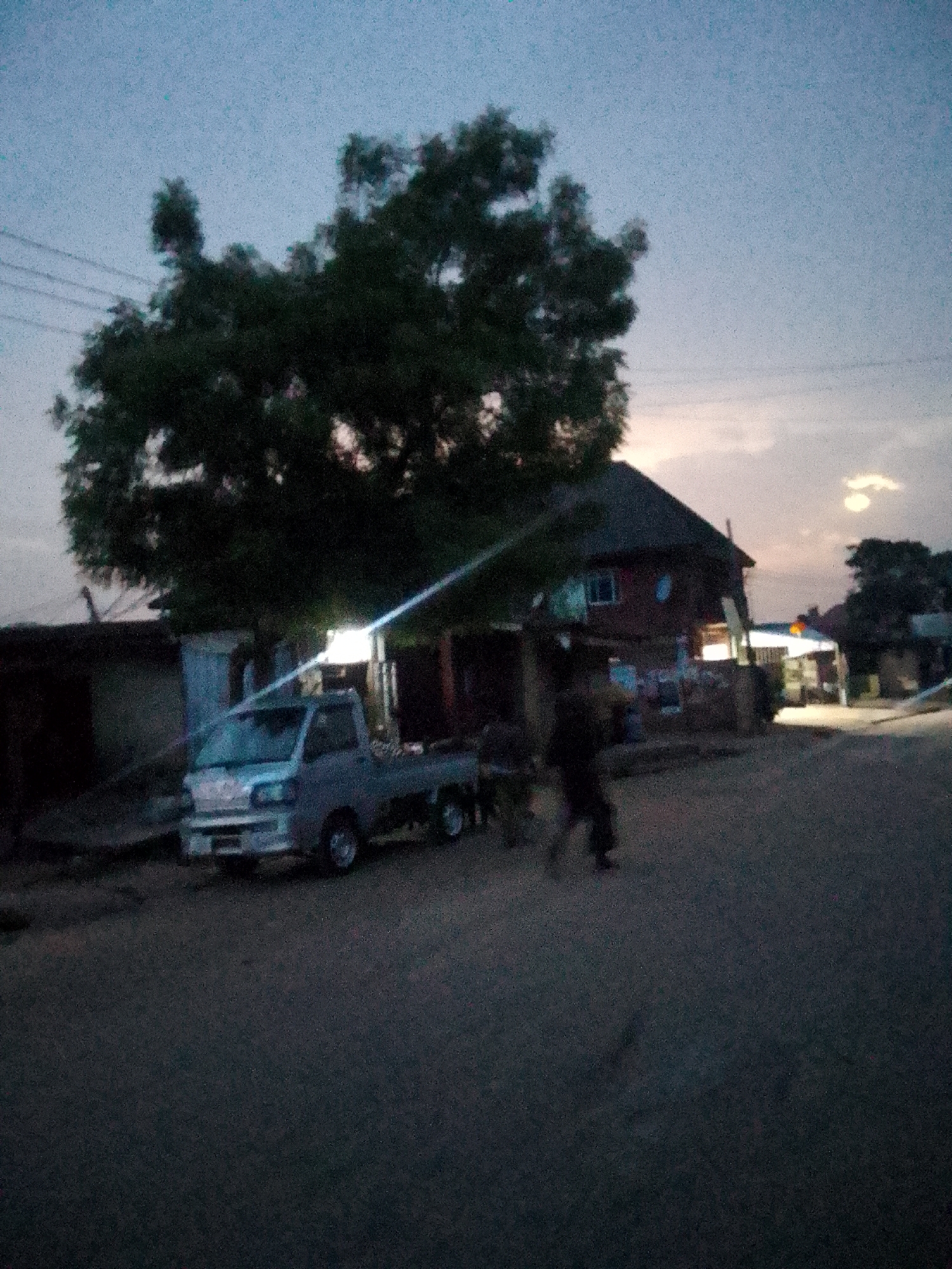 The image depicts a street scene in Onitsha, Nigeria, at dusk. A small, light-colored truck is parked near a building with an illuminated storefront. A large tree obscures part of the building. A person is walking on the street. Overhead wires are visible in the sky.