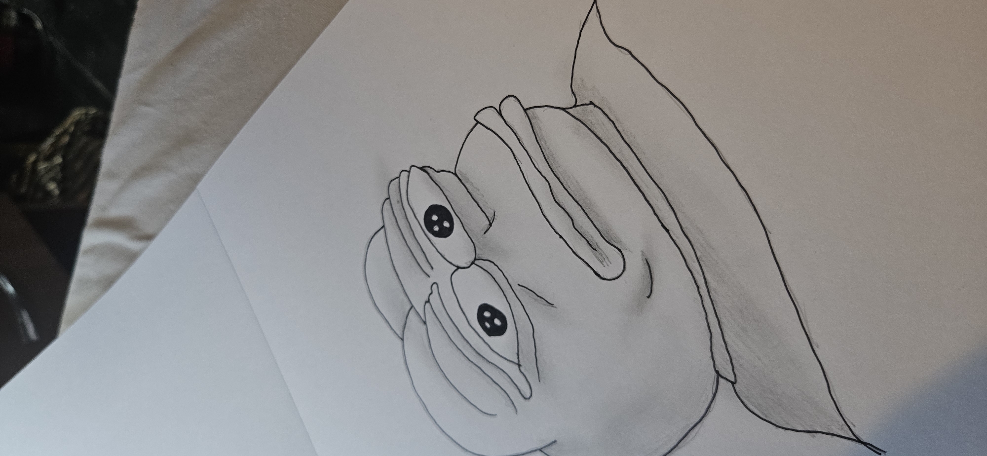 A pencil sketch of the Pepe the Frog meme is shown on a white piece of paper. The drawing is done in black and white and depicts the frog with its characteristic sad expression. The paper is lying on a textured surface, possibly a bed or a blanket, which is light brown in color. The background of the image is blurry and out of focus, showing a dark surface and some textured material.