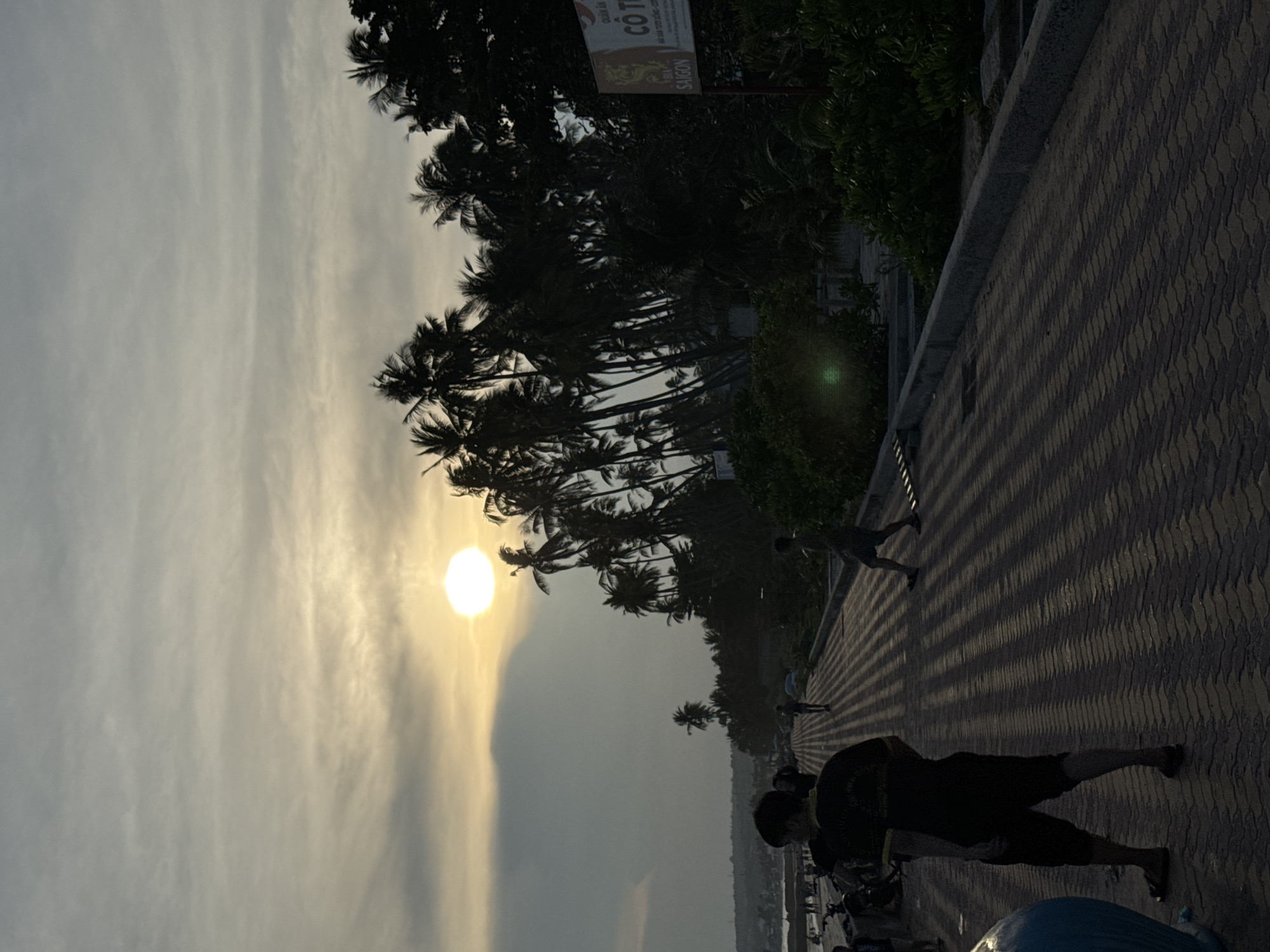 The image depicts a sunset scene on a paved walkway in Mũi Né, Vietnam. The sun, a bright orb, is partially obscured by clouds and is positioned in the upper left quadrant of the frame. A dense line of palm trees forms a silhouette against the dusky sky, extending across the right side of the image. The walkway, paved with patterned bricks, casts long, striped shadows due to the low angle of the sun.
In the foreground, on the left side of the walkway, a person wearing a dark t-shirt and shorts is walking away from the viewer, casting a prominent shadow. To the left of the walkway, a blue object, possibly a vehicle, is partially visible.
Further down the walkway, several other figures are present, silhouetted against the light. One individual is walking towards the sun, while another appears to be standing. In the background, on the left, a beach is visible with a scattering of people and what appear to be motorcycles parked. A sign with Vietnamese text and an emblem is mounted on a pole to the right, near the palm trees. The overall atmosphere is serene, capturing the end of the day in a coastal town.
