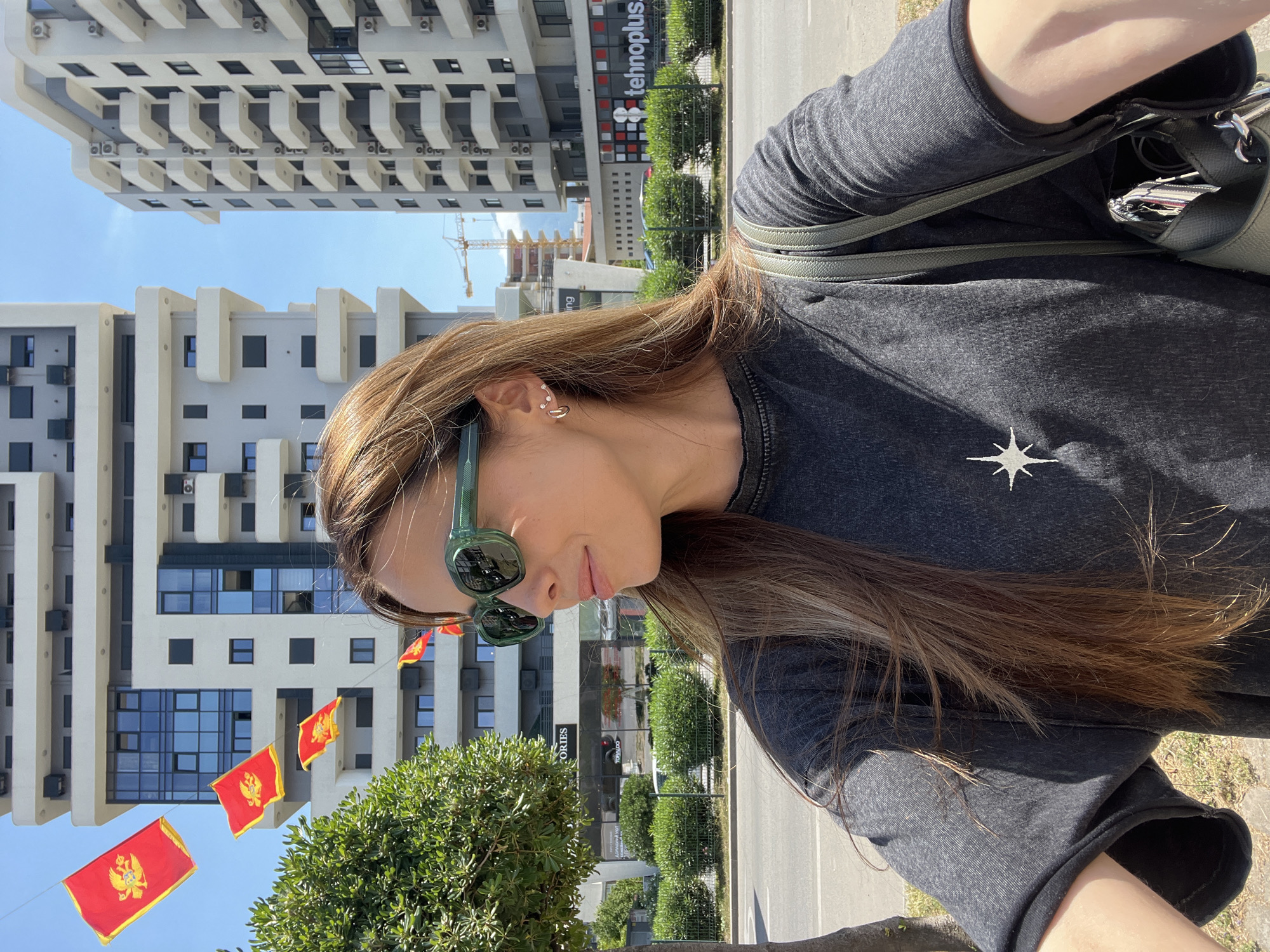 The image is a selfie taken outdoors in Bar, Montenegro. A woman with long, light brown hair and wearing green sunglasses and a dark gray t-shirt with a white star on the front is in the foreground. Behind her are several buildings, including a tall, modern apartment building with light-colored balconies and a sign reading "tehnoplus". There is also a building under construction with a visible crane. Montenegrin flags are strung between the buildings. Green trees and bushes line the street. The day appears sunny.