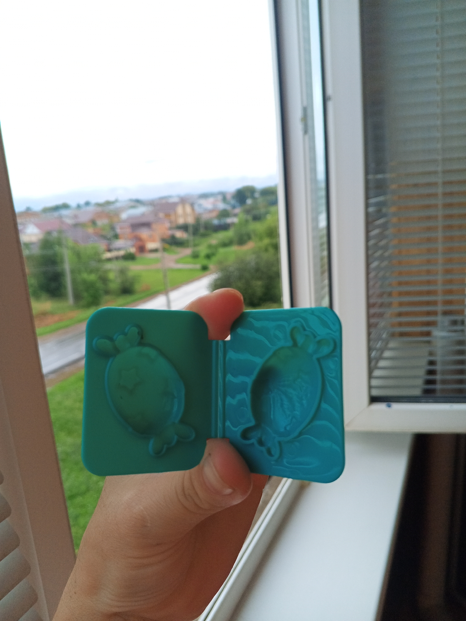 A human hand holds a turquoise plastic mold with two impressions of an oval shape with fins or decorative elements, set against a backdrop of a blurred cityscape visible through a window. The mold is open, revealing its two halves. The cityscape includes houses with red roofs, green trees, and a cloudy sky. To the right of the mold, a window blind with horizontal slats is visible. The lighting suggests it is daytime.