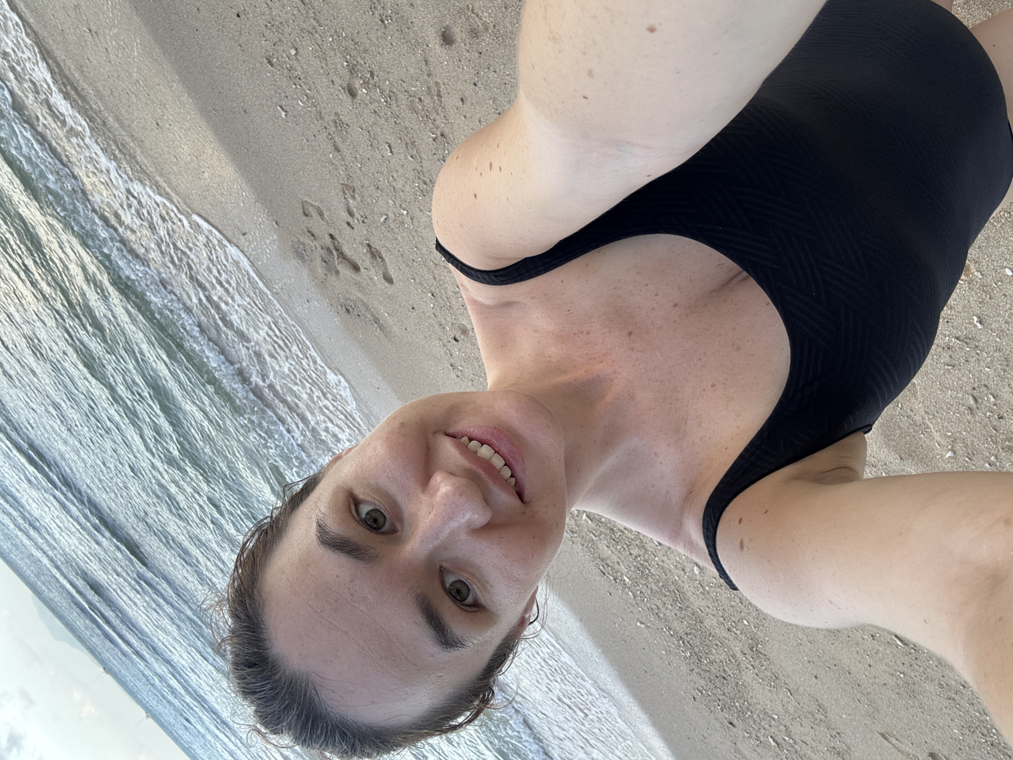 A woman in a black swimsuit takes a selfie on a sandy beach in Rawai, Thailand. The ocean waves gently lap the shore behind her. The sky is overcast, and a distant landmass is visible on the horizon. The woman is smiling, and her face is turned towards the camera. The sand is light brown and speckled with small shells. Footprints are visible in the sand.
