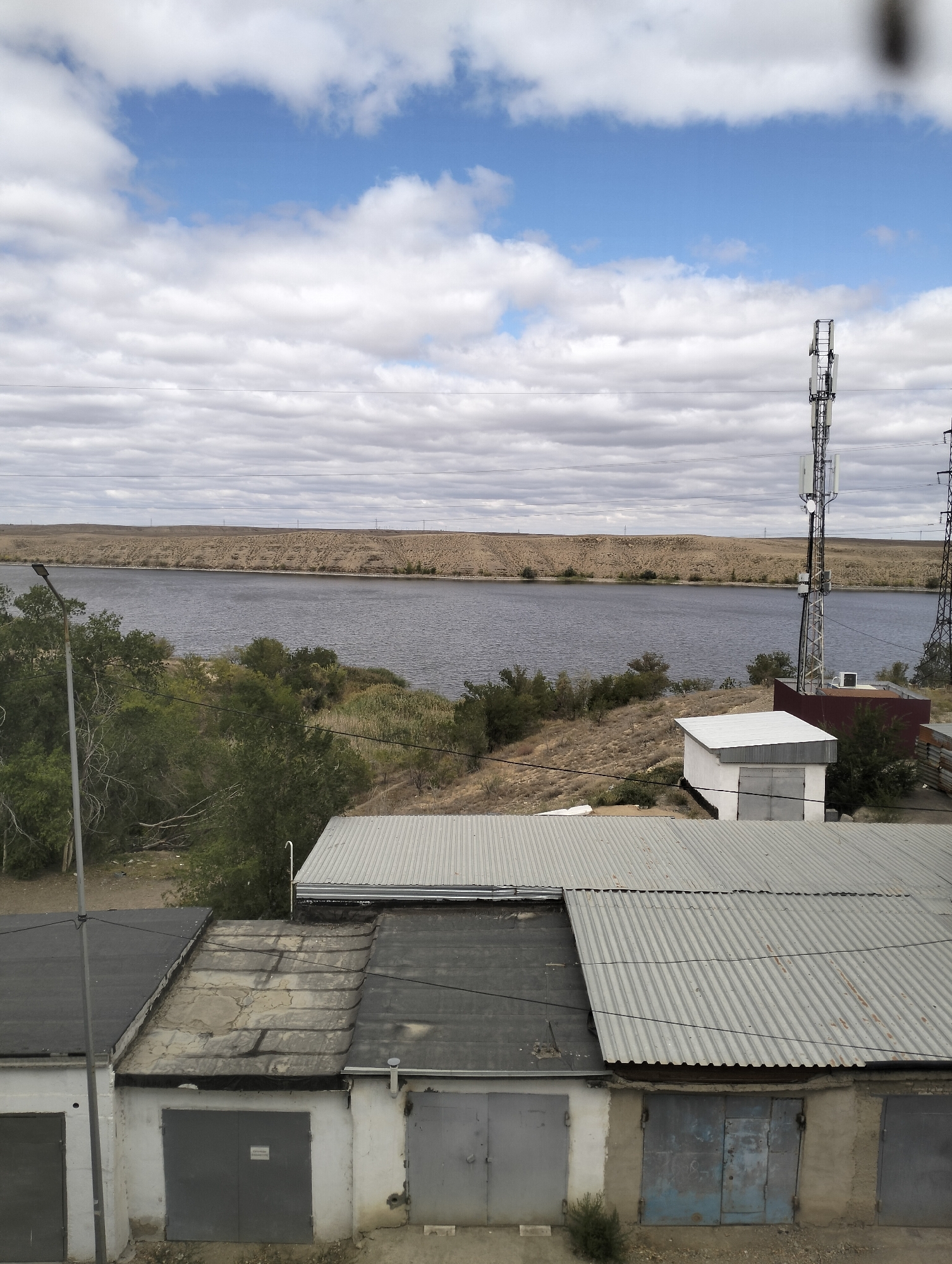 The image shows a panoramic view of a river landscape with a telecommunication tower and a row of garages in the foreground. The river is wide and calm, reflecting the cloudy sky. The opposite bank of the river is a dry, arid landscape with sparse vegetation. In the foreground, a metal telecommunication tower with multiple antennas stands tall. Below it, a small white building and a larger dark red structure are visible. A row of garages with corrugated metal roofs and large, grey doors occupies the bottom of the image. Power lines crisscross the scene. The sky is partly cloudy with patches of blue sky showing through. The overall impression is one of an industrial or semi-industrial area adjacent to a natural landscape. The specific location in Jezqazğan, Kazakhstan, suggests a region characterized by arid climate and industrial development.