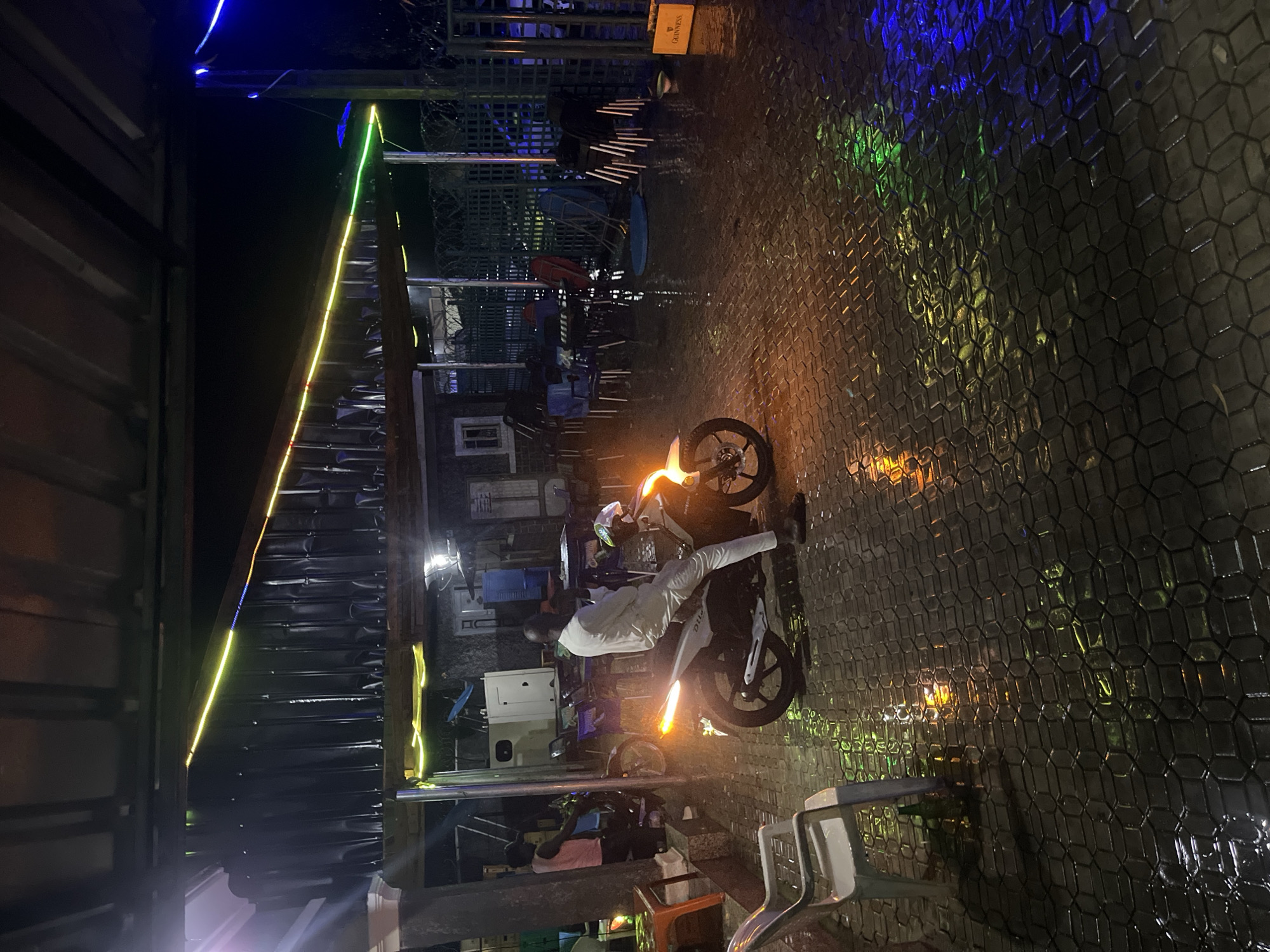 The image captures an outdoor nighttime scene in Jalingo, Nigeria, depicting a paved area with interlocking hexagonal blocks that are wet and highly reflective.
In the foreground and central mid-ground, a male subject dressed in a light-colored, possibly white, long-sleeved garment and dark sandals is seated stationary on a white scooter-style motorcycle. The motorcycle's rear light is illuminated orange, casting a distinct reflection on the wet ground directly beneath it.
To the left of the central motorcycle, another male subject wearing a light-colored, possibly pink or peach, short-sleeved top and dark trousers stands beside a darker two-wheeled vehicle, which appears to be a motorcycle or bicycle.
The overhead structure features a corrugated metal roof adorned with linear string lights that display segments of yellow, green, and blue illumination. Below this roofline, a building with a white facade and a small window is partially visible.
The scene is furnished with multiple plastic chairs of various colors, including white, blue, red, and black, some arranged around tables, others stacked or positioned on their sides. A white plastic chair is prominent in the immediate left foreground, with a dark green bottle positioned on the ground behind its front leg.
In the mid-background, a large white generator unit is present. The perimeter of the area is defined by a tall dark green fence topped with barbed wire, behind which additional structures are discernible. On the far right of the image, a wooden crate bearing the "GUINNESS" logo is placed on the wet pavement, adjacent to a small white bowl. The artificial light sources, including the string lights and motorcycle lights, cast multiple colorful reflections across the entire wet paved surface.