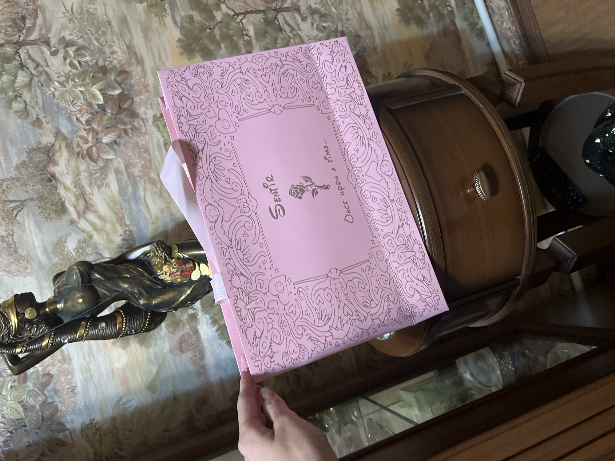 The image shows a pink box with a decorative pattern and the text "Sentir" and "Once upon a time..." in cursive font. The box appears to be a gift or product packaging. A bronze statue of a woman is partially visible on the left, lying down and adorned with gold accents. In the background, a wooden cabinet with a drawer is present, and to its right, a dark, round object with a sequined surface is visible. The setting appears to be indoors, with patterned wallpaper on the wall behind the statue. The lighting suggests it could be daytime, and the overall mood is serene.