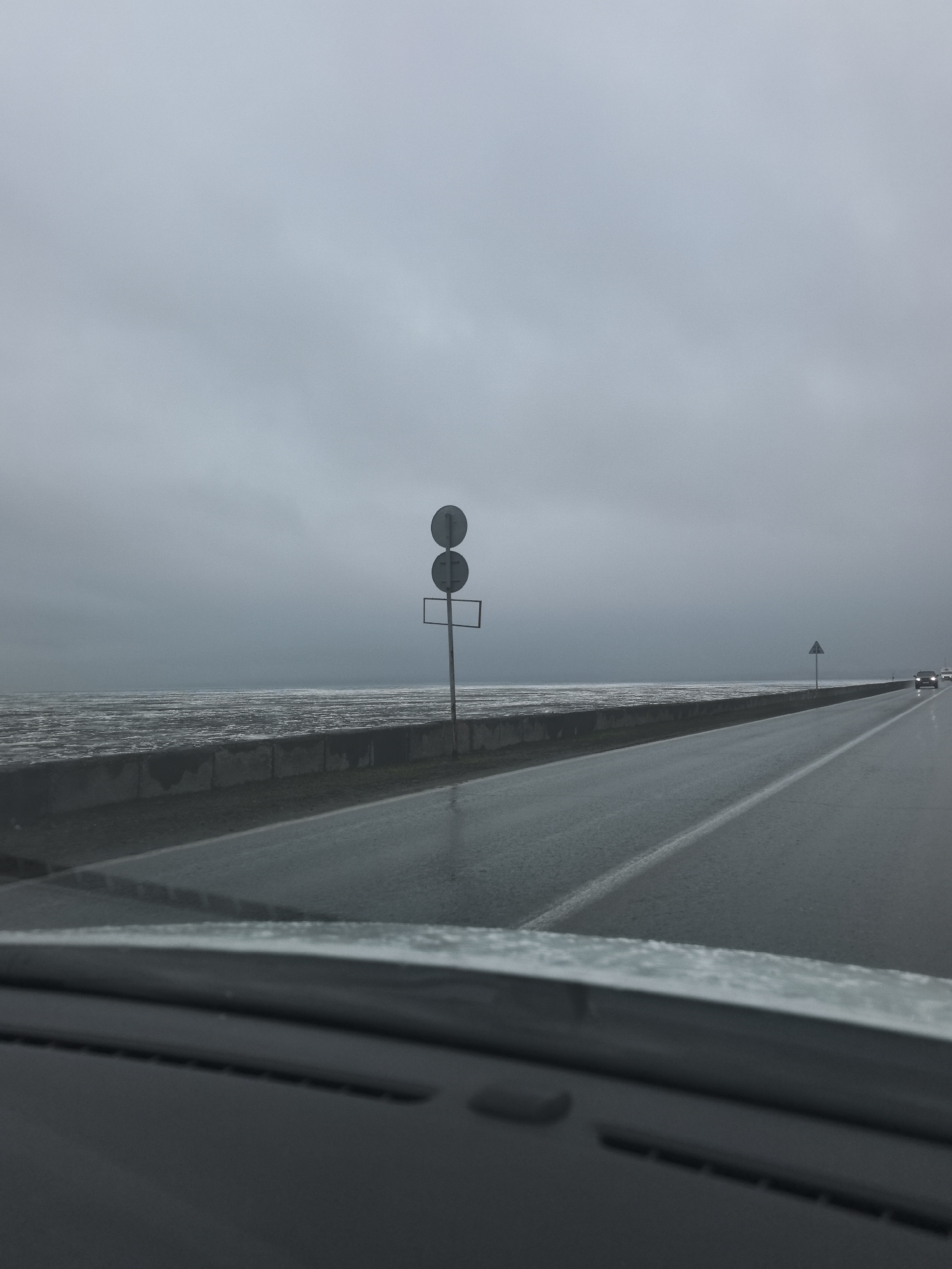 The image captures a road scene from the perspective of a vehicle's dashboard, likely in the Noviy region of Russia. A wet, two-lane highway stretches into the distance under a uniformly overcast, grey sky.
On the left, the road is bordered by a concrete barrier that separates it from a vast body of water, possibly a sea or bay. The water's surface appears dark, turbulent, and highly reflective, or potentially covered in ice floes, blending indistinctly with the horizon line.
Mid-ground on the left, a tall metal post displays two circular traffic signs mounted vertically, with an empty rectangular sign panel below them. Their specific meaning is not discernible due to distance. Further down the road on the right, a triangular warning sign with a dark border is partially visible.
The road surface is dark and visibly wet, suggesting recent or ongoing rain. In the far distance on the right lane, the illuminated headlights of at least two approaching vehicles can be seen. The foreground shows the dark dashboard and the lower part of the windshield, which has visible water droplets, confirming the wet weather.
The overall atmosphere is one of a bleak, grey, and possibly cold day. There is no discernible text visible in the image.