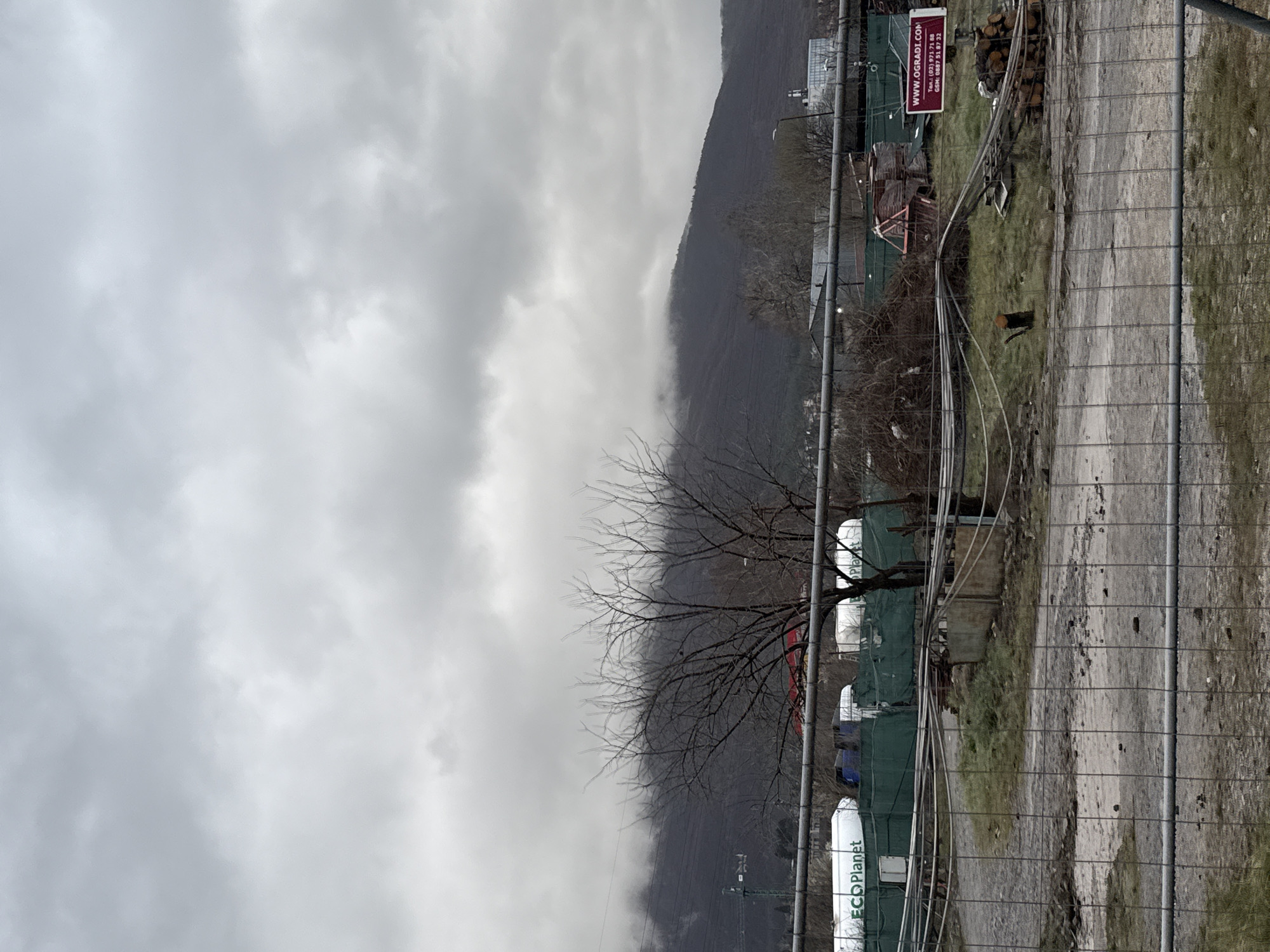 The image shows a scene in Sofia, Bulgaria on an overcast day. A mountain is partially obscured by low-hanging clouds. In the foreground, there's a construction or industrial area demarcated by a chain-link fence. Within the fenced area are storage tanks with "ECO Planet" printed on them. A bare tree stands near the tanks. A sign with "WWW.OGRADI.COM" and Bulgarian telephone numbers is visible, along with a pile of logs. The ground appears muddy and uneven.