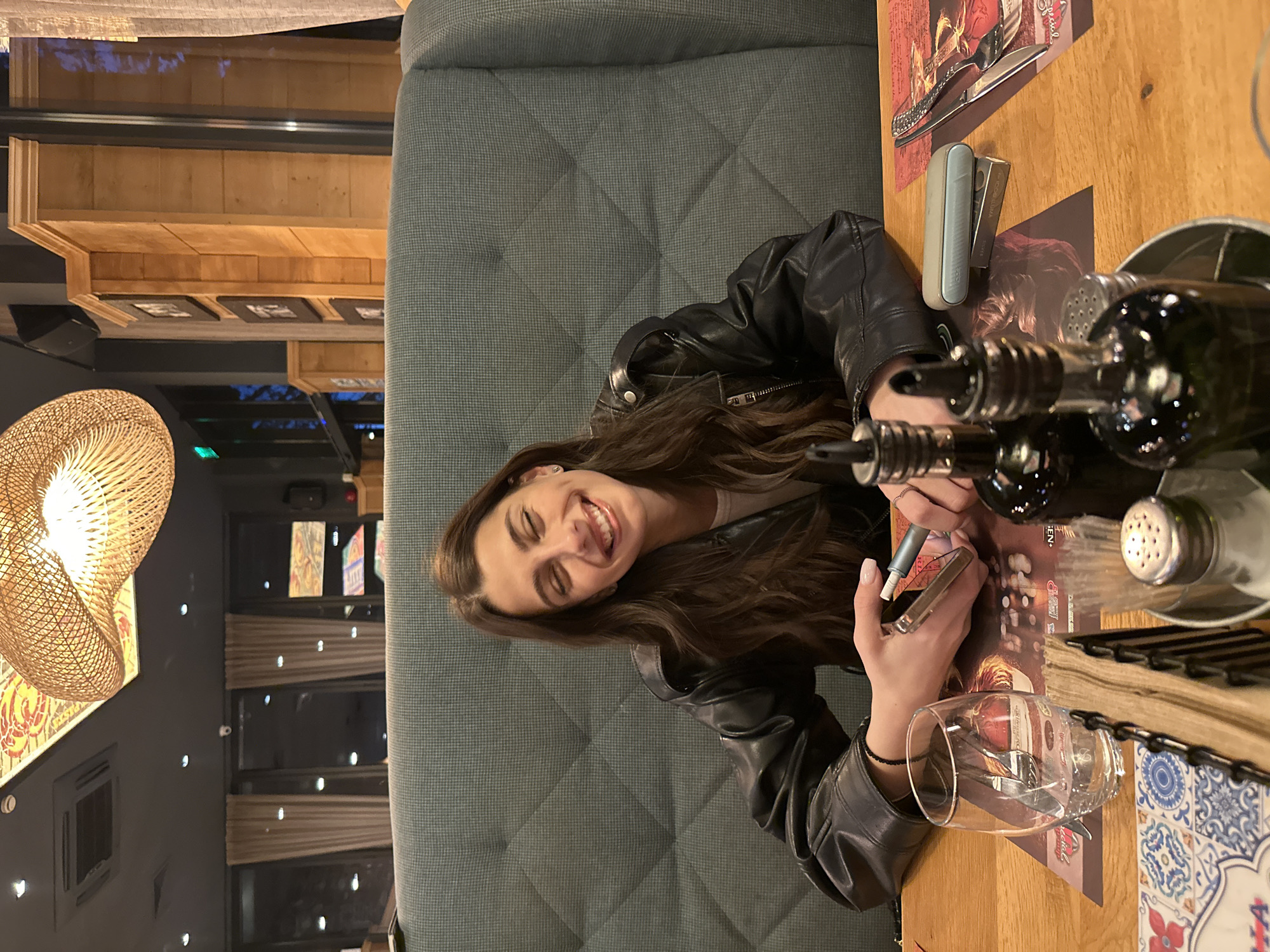 An indoor scene in what appears to be a restaurant or cafe in Plovdiv, Bulgaria, captures a young woman smiling broadly at the camera. The image is oriented sideways, with the woman seated in a dark grey upholstered booth on the left, at a light-colored wooden table. She has long, dark, wavy hair and is wearing a black leather jacket.
Her right hand holds a pen or marker, actively writing or drawing on a paper placemat or menu. Her left hand holds a smartphone. On the table, to her right, there is an oil or vinegar dispenser set, a salt shaker, a clear drinking glass, and cutlery. A light blue and silver IQOS heated tobacco device is also visible on the table. The placemat features decorative patterns and some partially visible text, including the letters "MEN".
The background reveals more of the establishment's interior, including a large, woven, spherical light fixture casting a warm glow, dark walls with decorative accents, and wooden wall panels. Window blinds are visible in the distance. The woman's joyful expression and the casual dining setting suggest a relaxed and pleasant atmosphere, possibly during an evening outing.