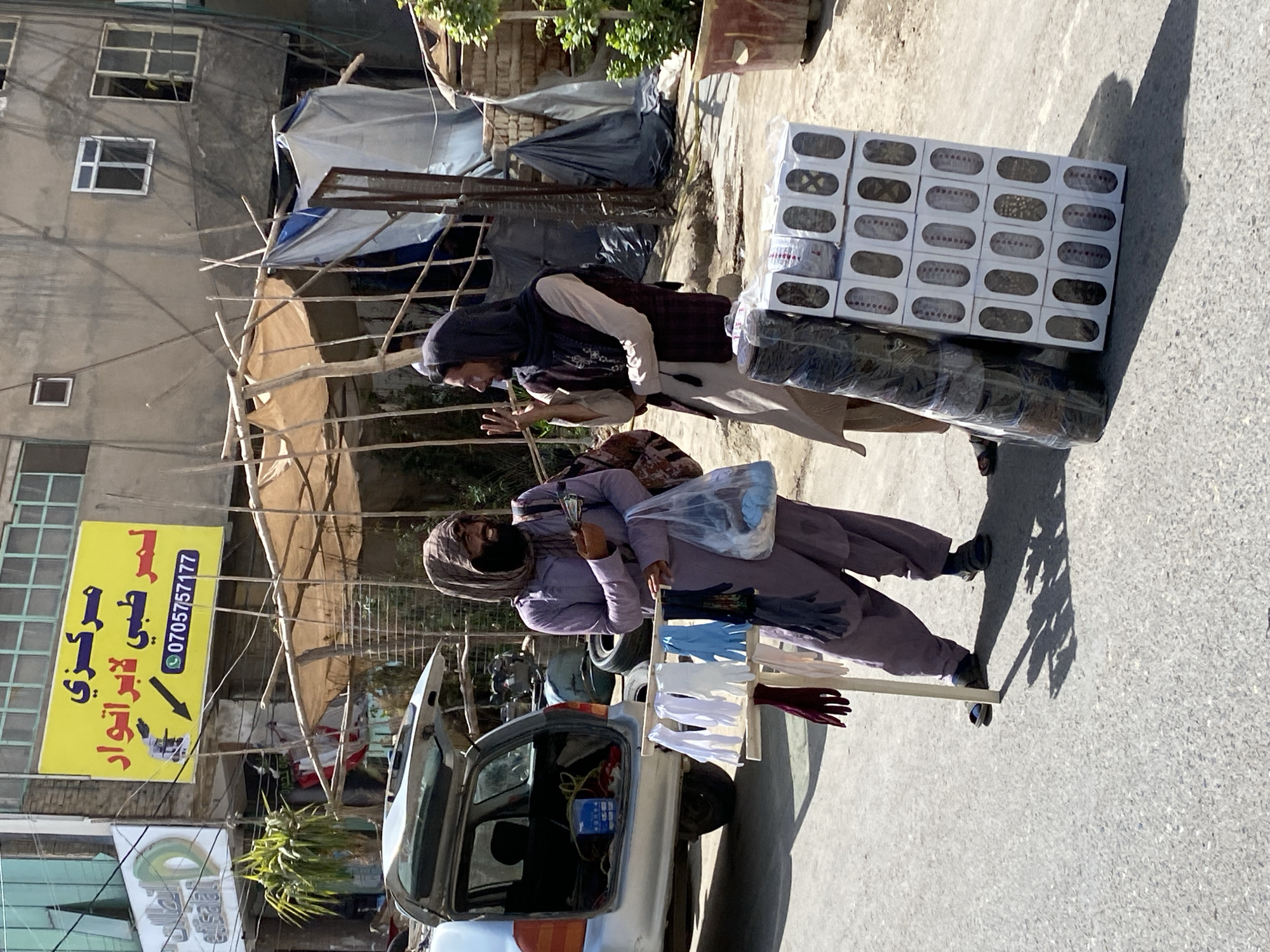 Two men are standing on a paved street in front of a building in Kandahar, Afghanistan. The man on the left is wearing a purple tunic and pants and a headscarf. He is holding a plastic bag in his left hand and appears to be talking and gesturing with his right hand. In front of him is a wooden rack displaying several pairs of gloves in various colors. To his right is a silver car with its trunk open, containing some items.
The man on the right is wearing a dark vest over a light-colored tunic and pants, and a dark headscarf. He is holding his right hand up, with fingers extended, and appears to be speaking to the other man. To his right is a stack of white boxes, each containing what appear to be slippers or shoes, wrapped in clear plastic. Another larger cylindrical object wrapped in plastic is leaning against the boxes.
The background shows a building with a yellow sign that has Arabic writing and a phone number. There are some temporary structures with fabric coverings, as well as a potted plant. The scene appears to be an outdoor market or street vendor area. The lighting suggests it is daytime, with distinct shadows cast on the ground.