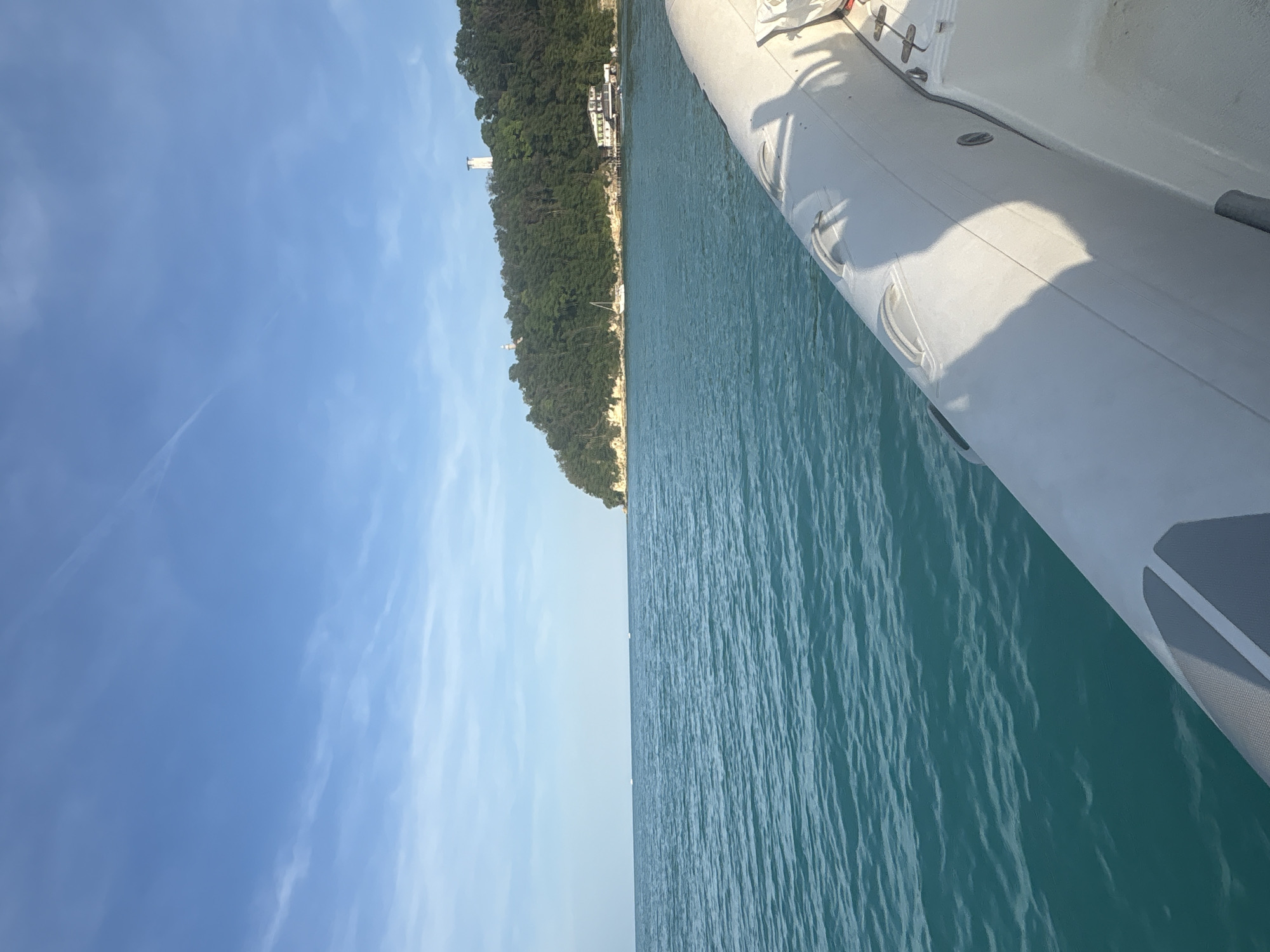 The image depicts a daytime maritime scene captured from the starboard side of a boat. In the immediate foreground, the white inflatable pontoon of a Rigid Inflatable Boat (RIB) occupies the lower right quadrant, displaying multiple integrated handles and a prominent shadow cast upon its surface. Adjacent to the pontoon, a section of the boat's white deck with grey textured elements is visible.
The midground features a vast expanse of turquoise-to-teal colored water, exhibiting a textured surface with small ripples. The water extends to the horizon on the left, where two faint white specks, possibly distant boats, are discernible.
To the right, a densely forested landmass, likely an island or coastline, rises from the water. Its base reveals exposed light-colored rock formations at the waterline. Perched atop the forested hill are two distinct white tower-like structures; one is a slender column, and the other is a broader, multi-level tower, both resembling lighthouses or navigational aids. Below these towers, near the shore, a multi-story building with a white and green exterior is visible, partially obscured by trees. A white sailboat is anchored near the shoreline, positioned to the left of the building.
The background consists of a clear, light blue sky with scattered thin, wispy clouds and a faint contrail stretching diagonally in the upper right. The lighting indicates a bright, sunny day, with shadows consistent with direct sunlight. The location is an unspecified coastal or island environment in Unknown, Unknown.